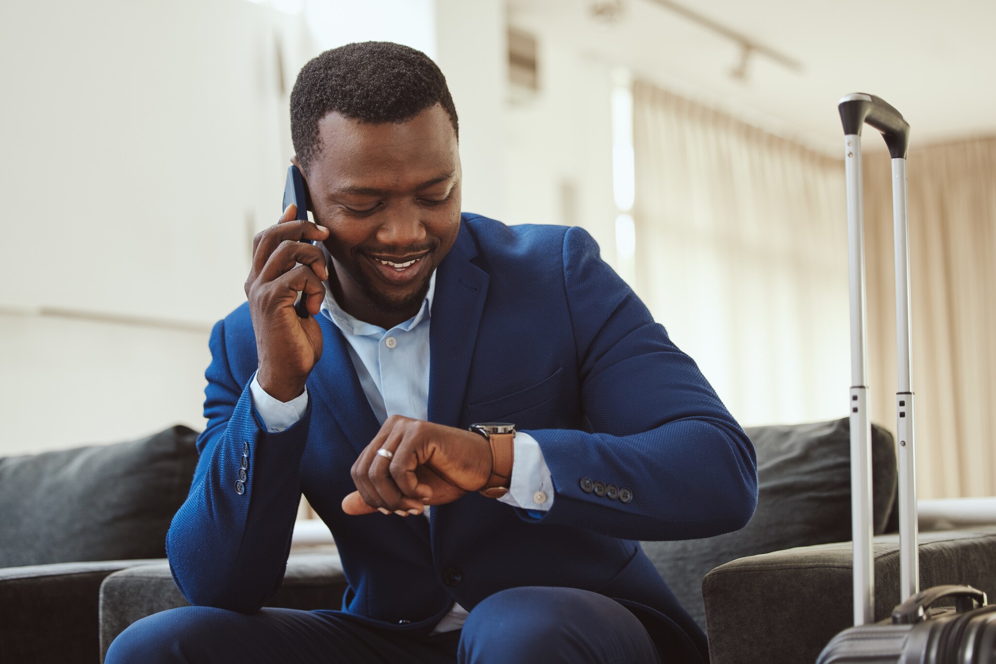 Ein lächelnder Geschäftsmann sitzt mit Smartphone am Ohr in einer Lounge und blickt auf seine Armbanduhr.
