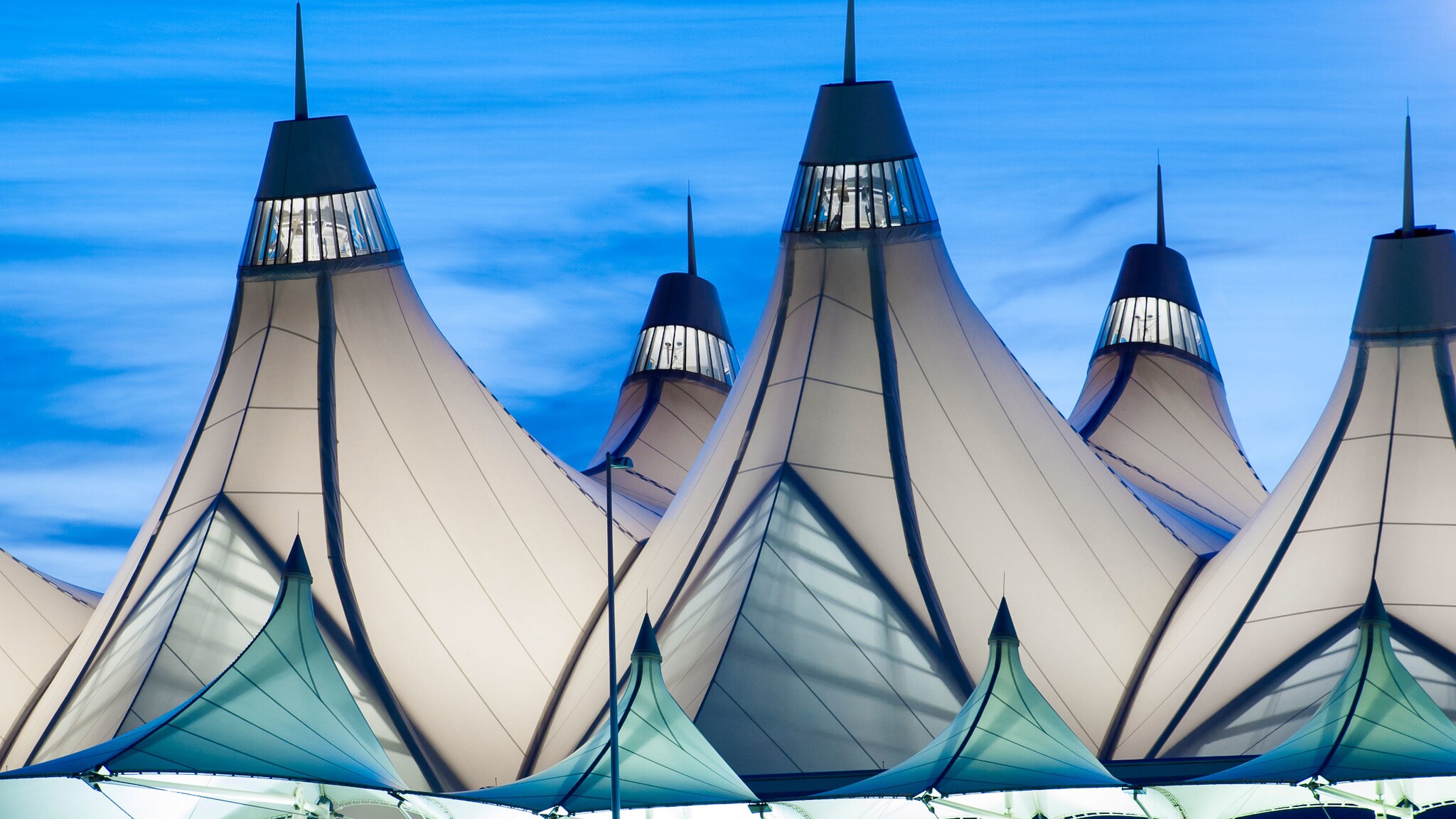 Beleuchtetes Gebäudedach aus Fibersegeltuch in konischen Formen vor blauem Himmel am Abend.