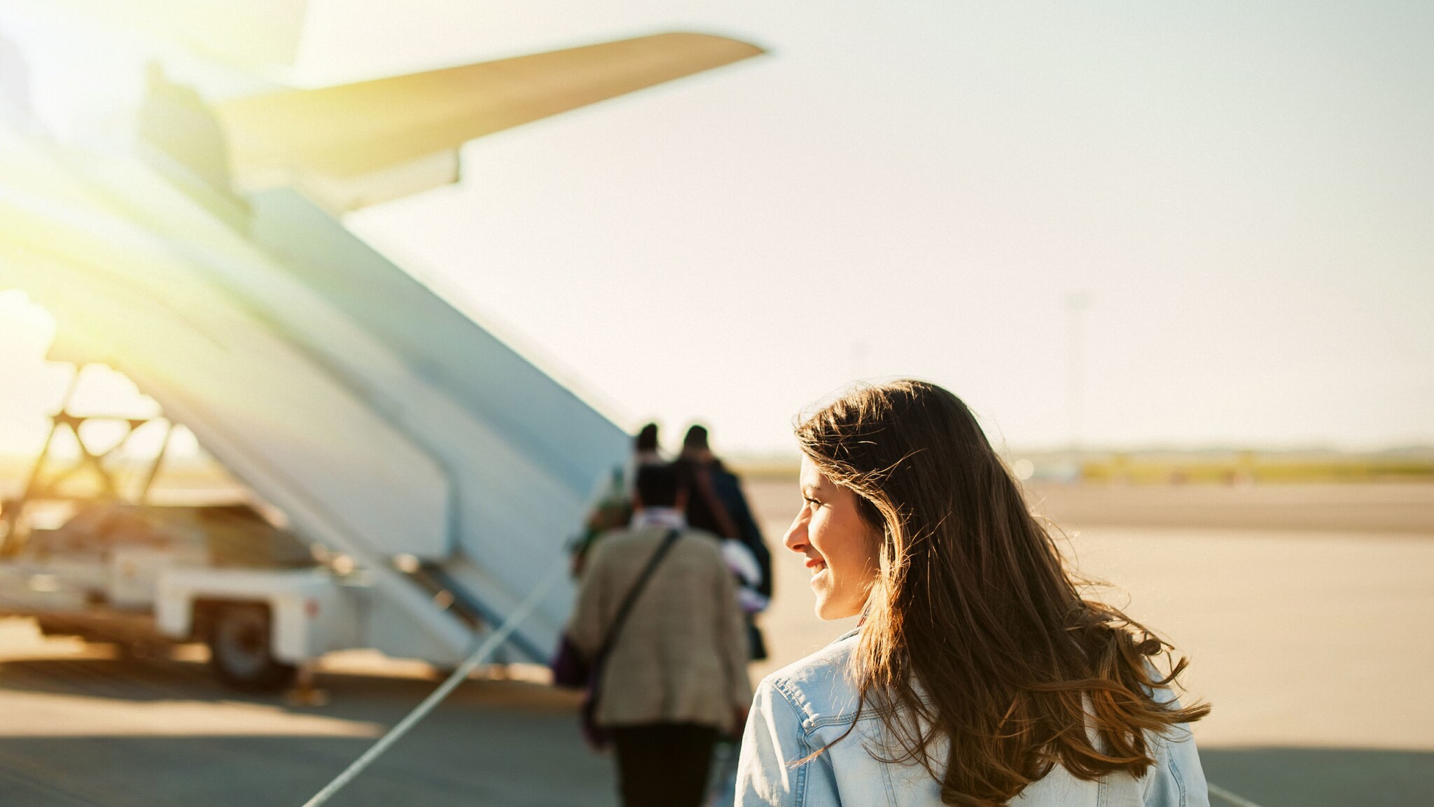 Eine Frau vor einem Flugzeug auf dem Rollfeld eines Flughafens, über dessen Treppe Passagiere einsteigen.
