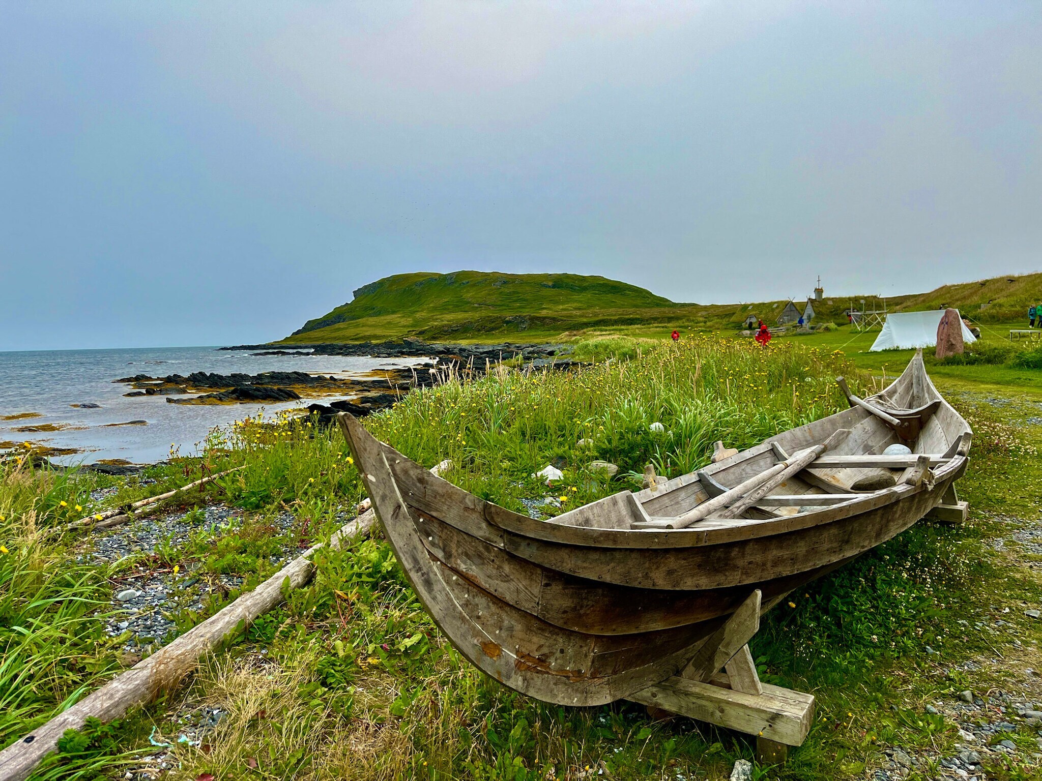 Das Bild zeigt ein altes Holzboot auf einer grünen Wiese an der Küste. Im Hintergrund sind ein Zelt und ein mit Gras bewachsenes Haus zu sehen.