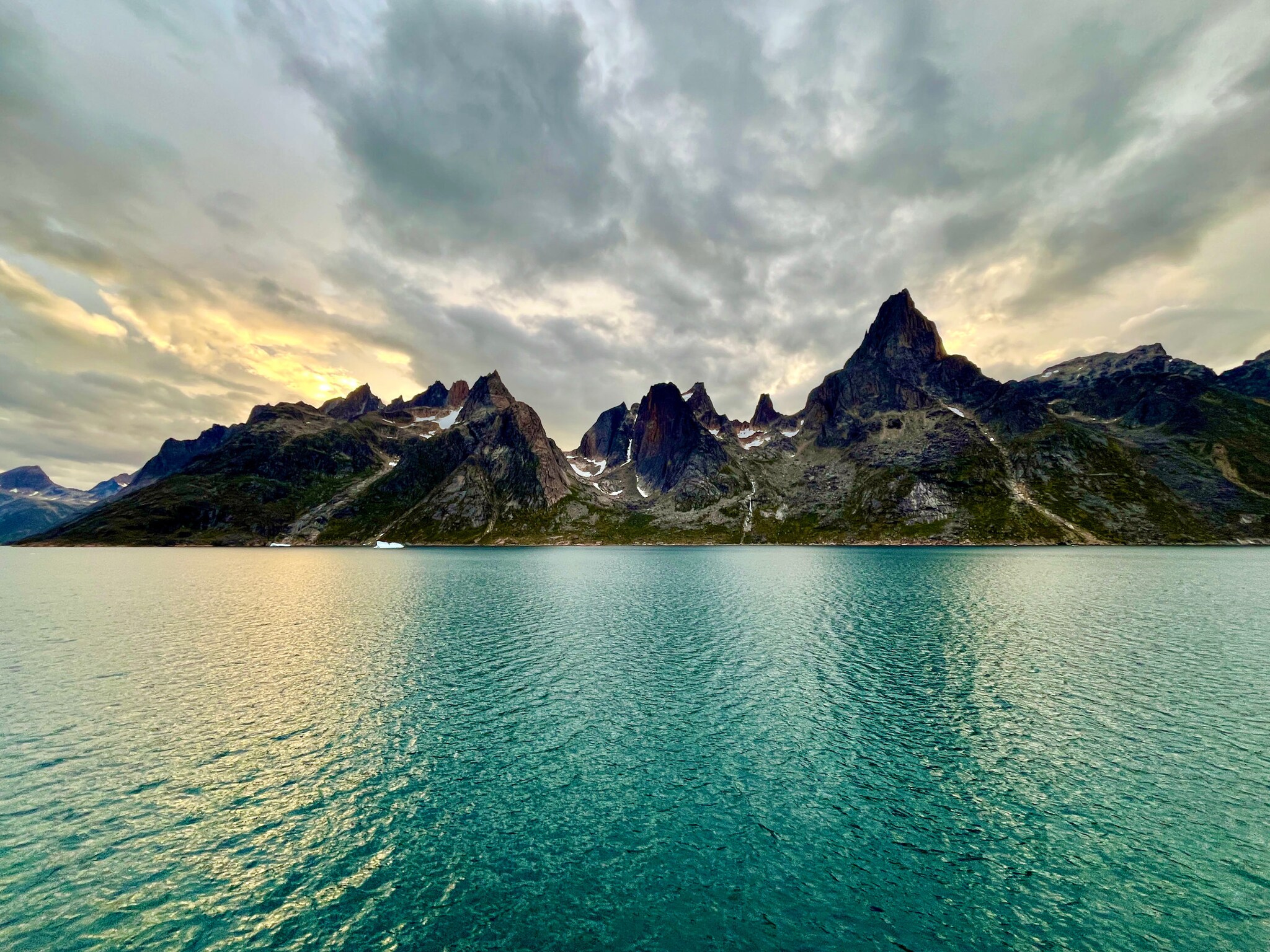 Das Bild zeigt hohe und spitze Berge. Im Vordergrund ist das grünlich schimmernde Wasser des Prins Christian Sunds zu sehen.
