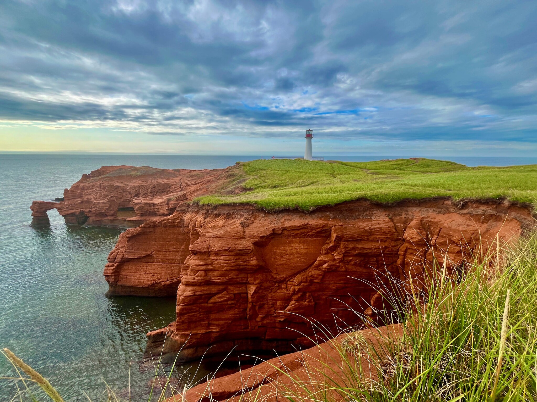 Das Bild zeigt die Steilküste im Osten von Kanada. Rote Felsen erheben sich aus dem Meer. Oberhalb sind hellgrünes Gras und ein weißer Leuchtturm zu sehen. Das Bild zeigt die Steilküste im Osten von Kanada. Rote Felsen erheben sich aus dem Meer. Oberhalb sind hellgrünes Gras und ein weißer Leuchtturm zu sehen.