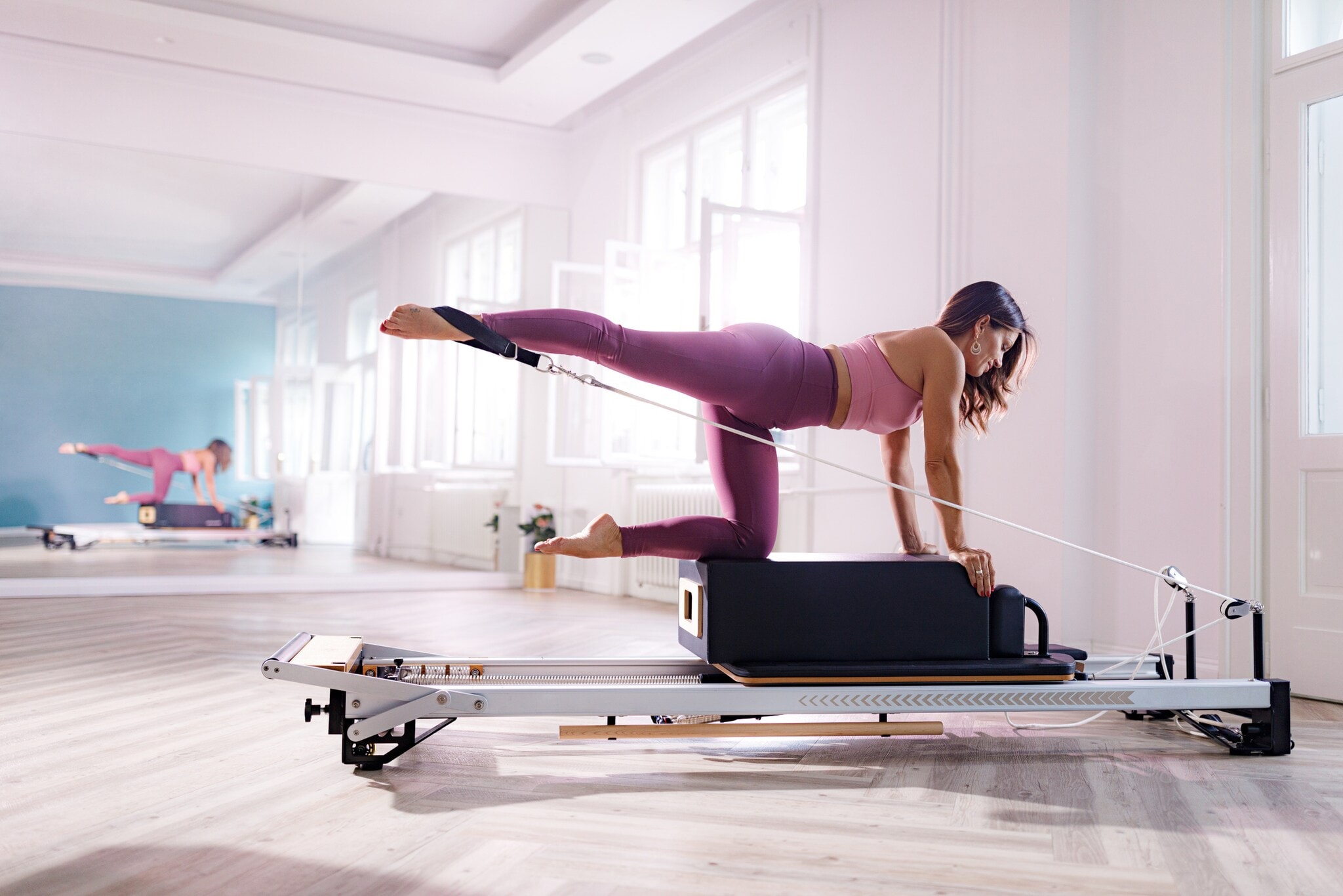 Eine Frau in pinker Sportkleidung kniet auf einer Box auf einem Pilates Reformer in einem hellen Raum. Eine Frau in pinker Sportkleidung kniet auf einer Box auf einem Pilates Reformer in einem hellen Raum.