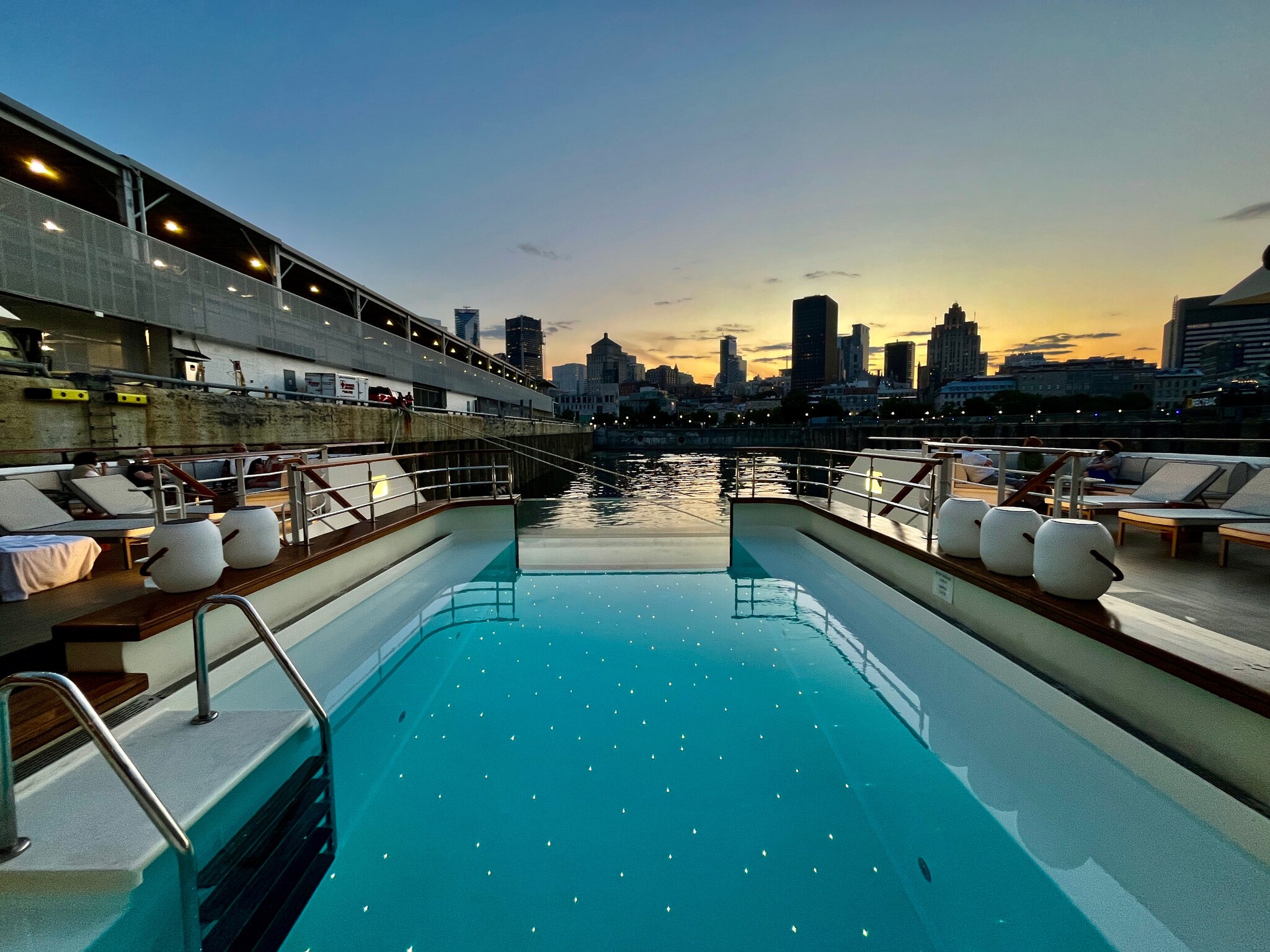 Das Bild zeigt einen Pool mit Liegen an Bord des Schiffes Le Bellot. Im Hintergrund sieht man die Umrisse der Hochhäuser der Skyline von Montreal. Dahinter geht die Sonne unter.