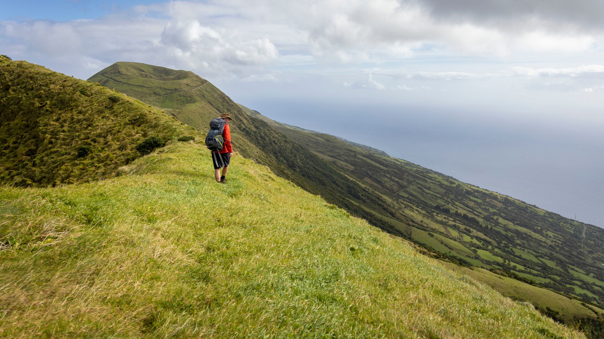 Eine Person mit Wanderausrüstung auf einem grün bewachsenen und steil abfallenden Vulkankegel der Azoreninsel São Jorge.