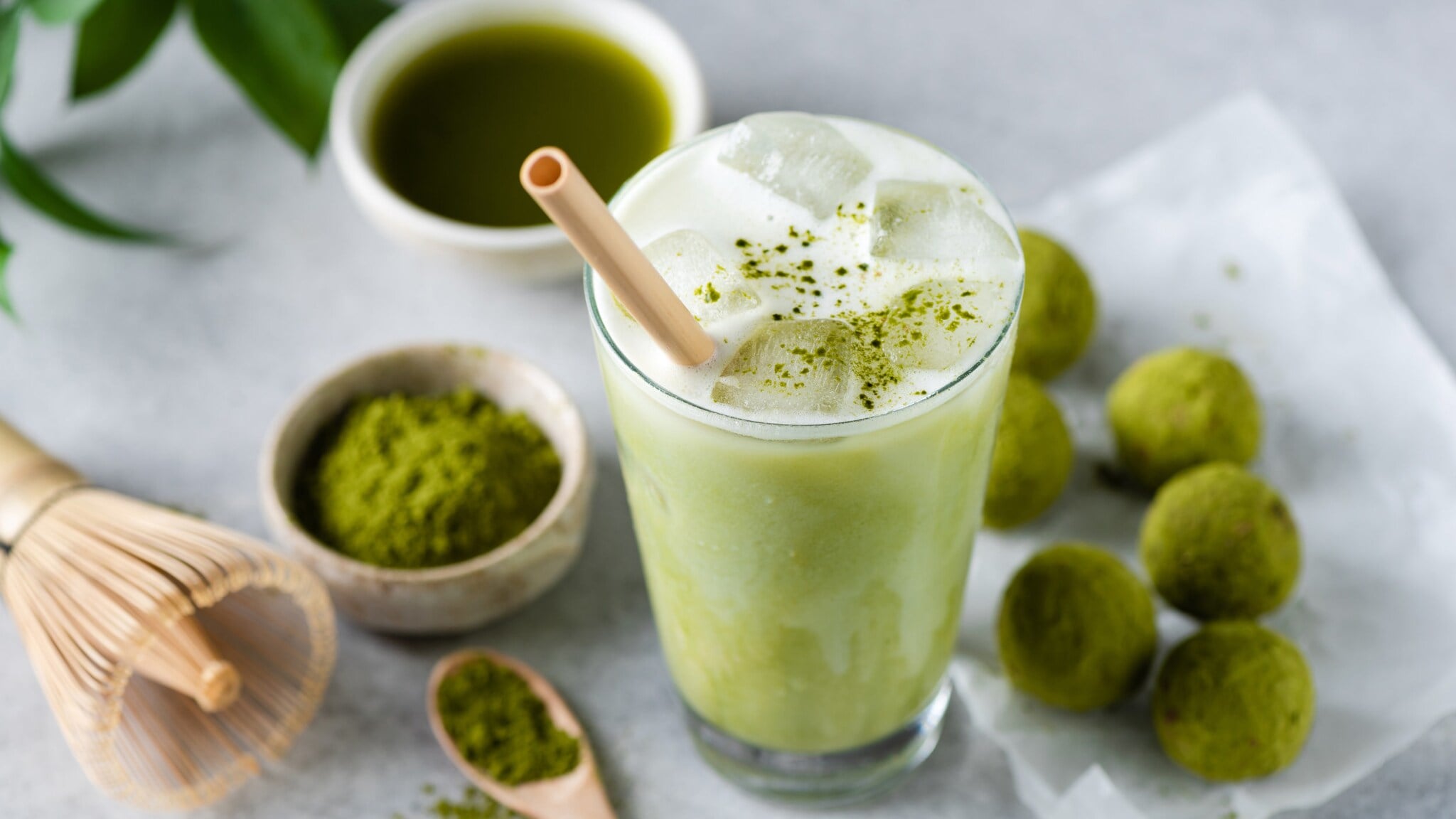 Ein Glas mit Matcha Latte und Strohhalm, daneben liegen grüne Kugeln, eine Schale mit grünem Pulver, ein Bambusbesen und eine Schale mit purem Matchatee. Ein Glas mit Matcha Latte und Strohhalm, daneben liegen grüne Kugeln, eine Schale mit grünem Pulver, ein Bambusbesen und eine Schale mit purem Matchatee.