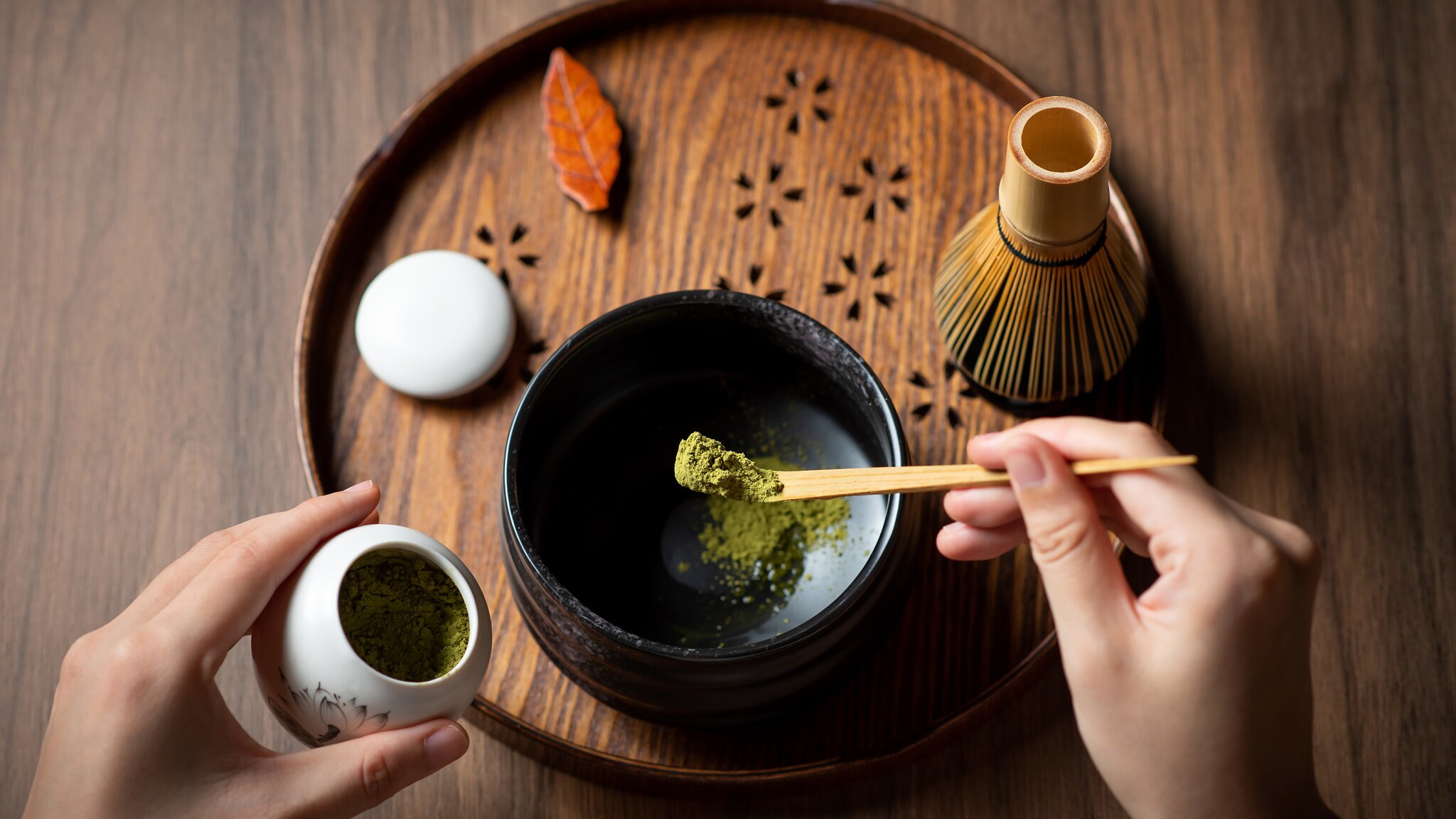 Eine unkenntliche Person gibt mit einem Holzlöffel etwas Matchapulver in eine Schüssel, die auf einem Tablett steht. Daneben befindet sich ein Bambusbesen.