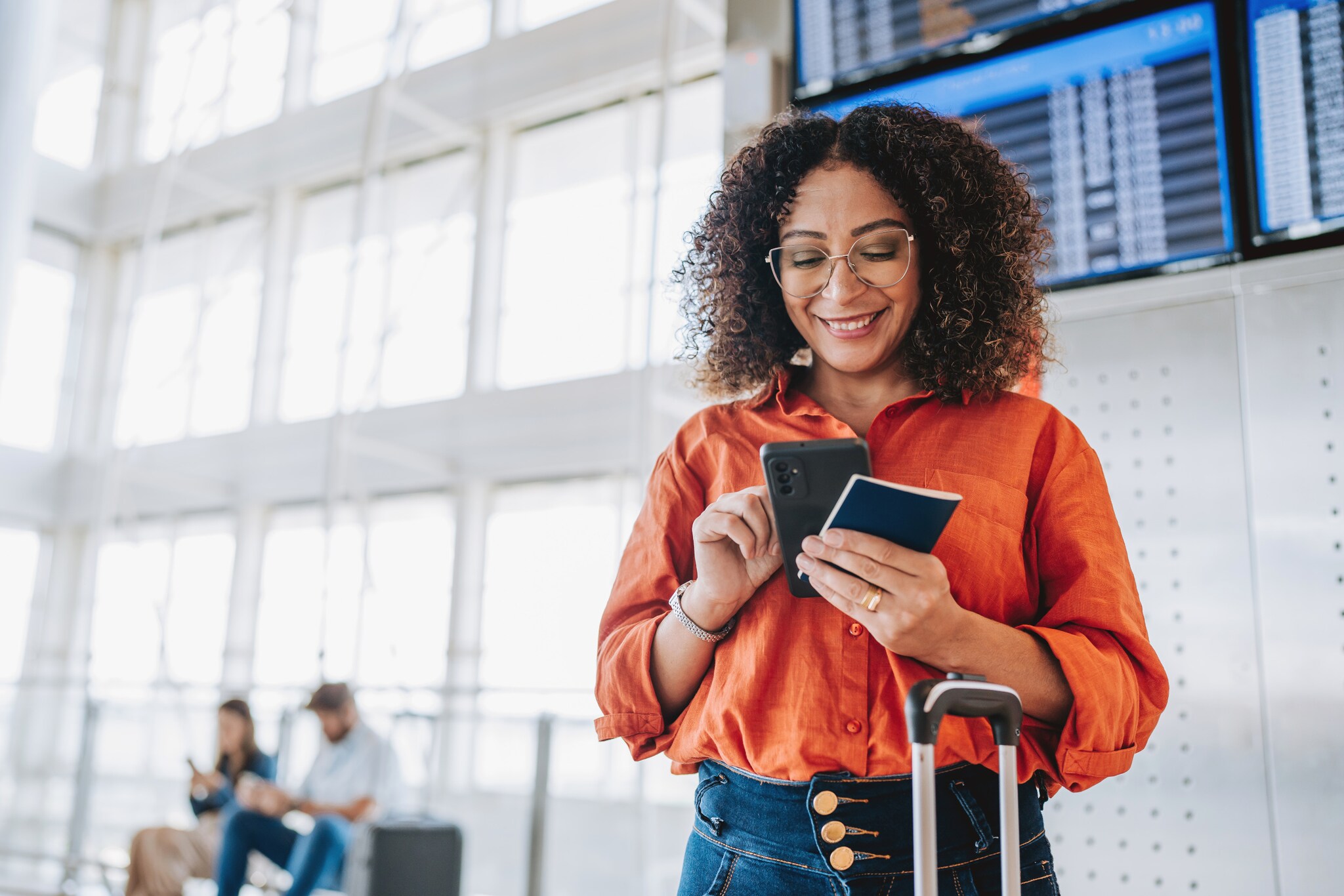 ine Frau bedient lächelnd ihr Smartphone in einem Flughafenterminal, in ihrer Hand ein Reisepass.