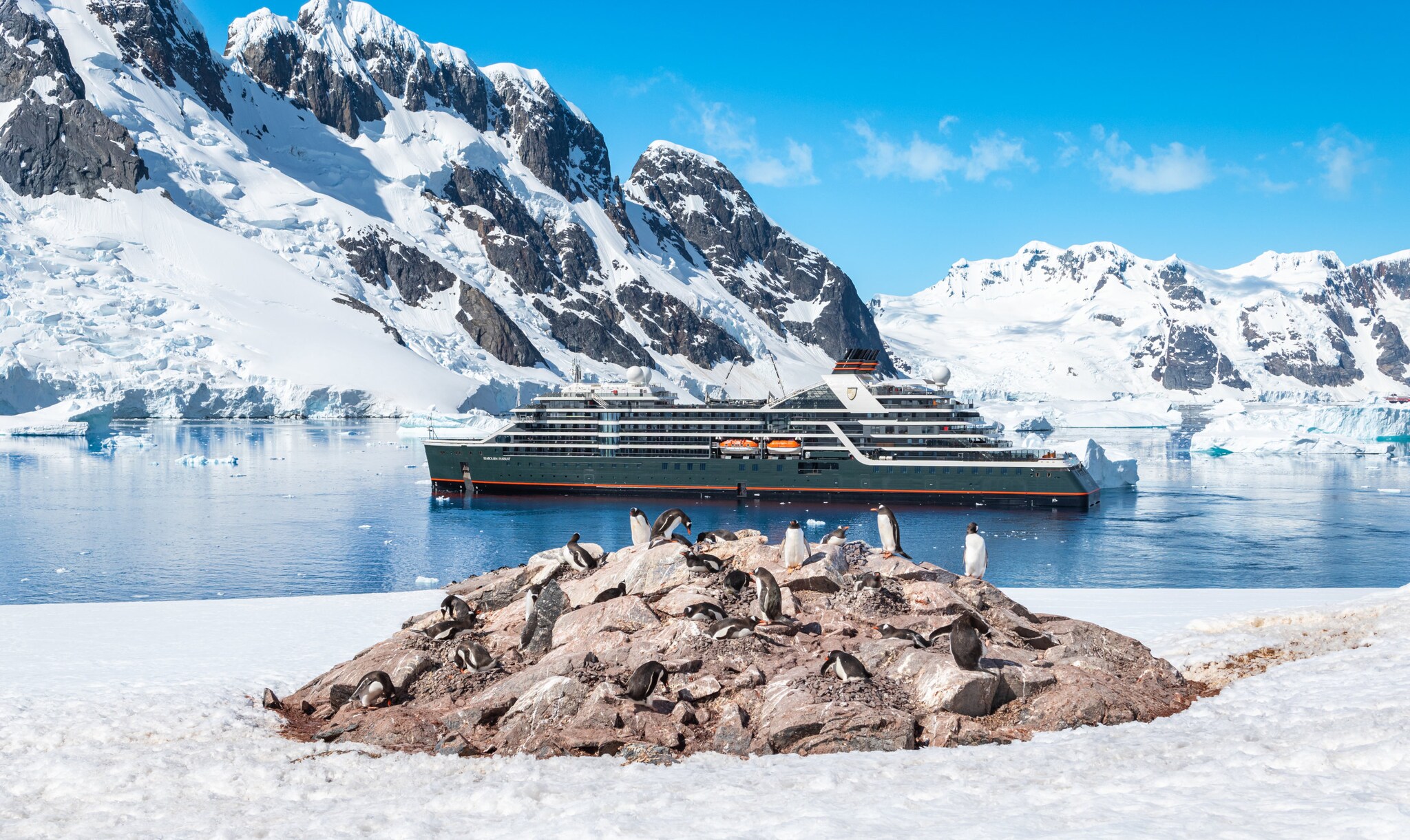 Ein Expeditionsschiff in der Antarktis, im Vordergrund Pinguine auf Felsbrocken im Schnee.