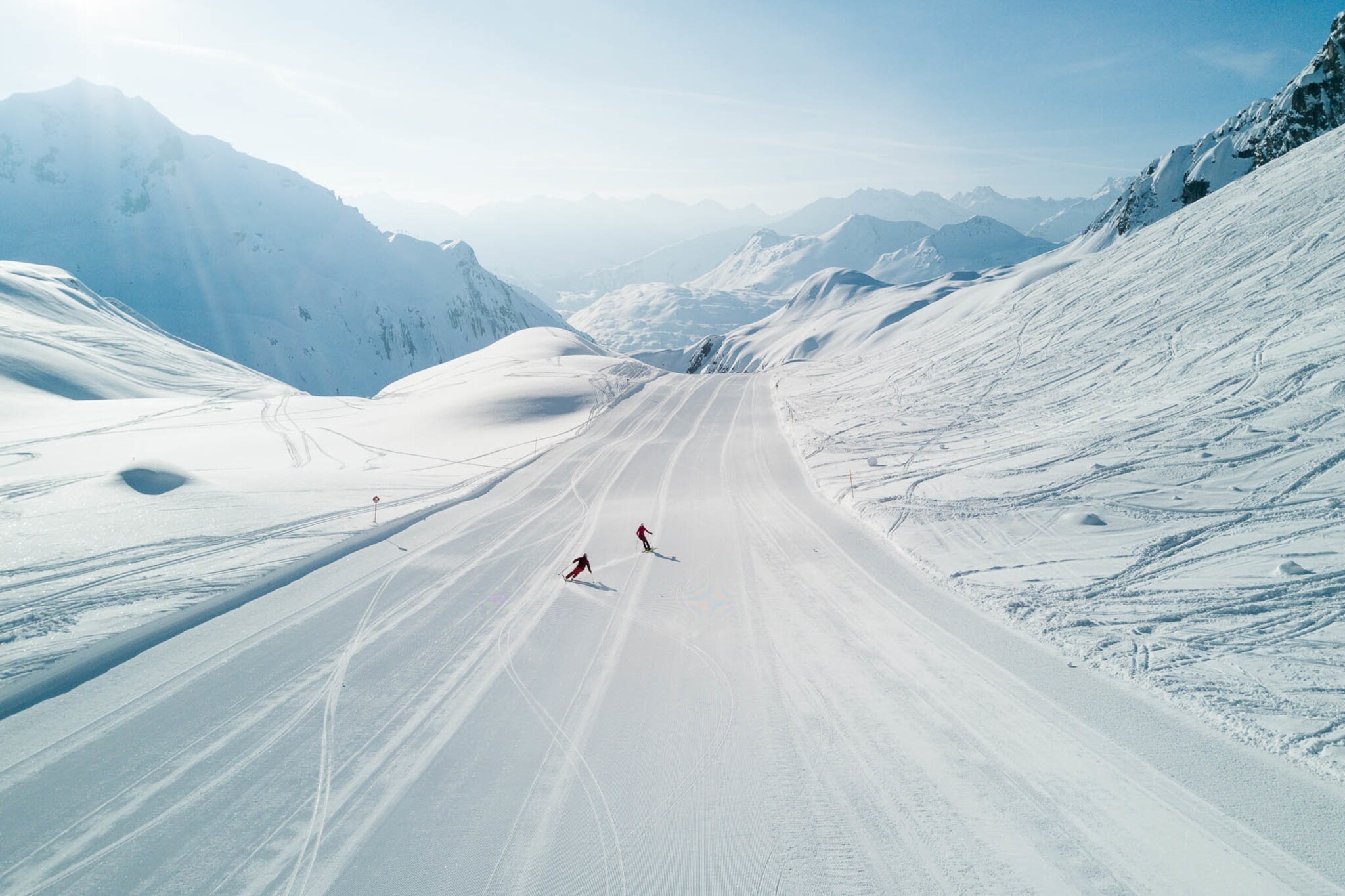 Zwei Skifahrende auf einer breiten Piste inmitten einer Schneelandschaft vor Gipfelpanorama.