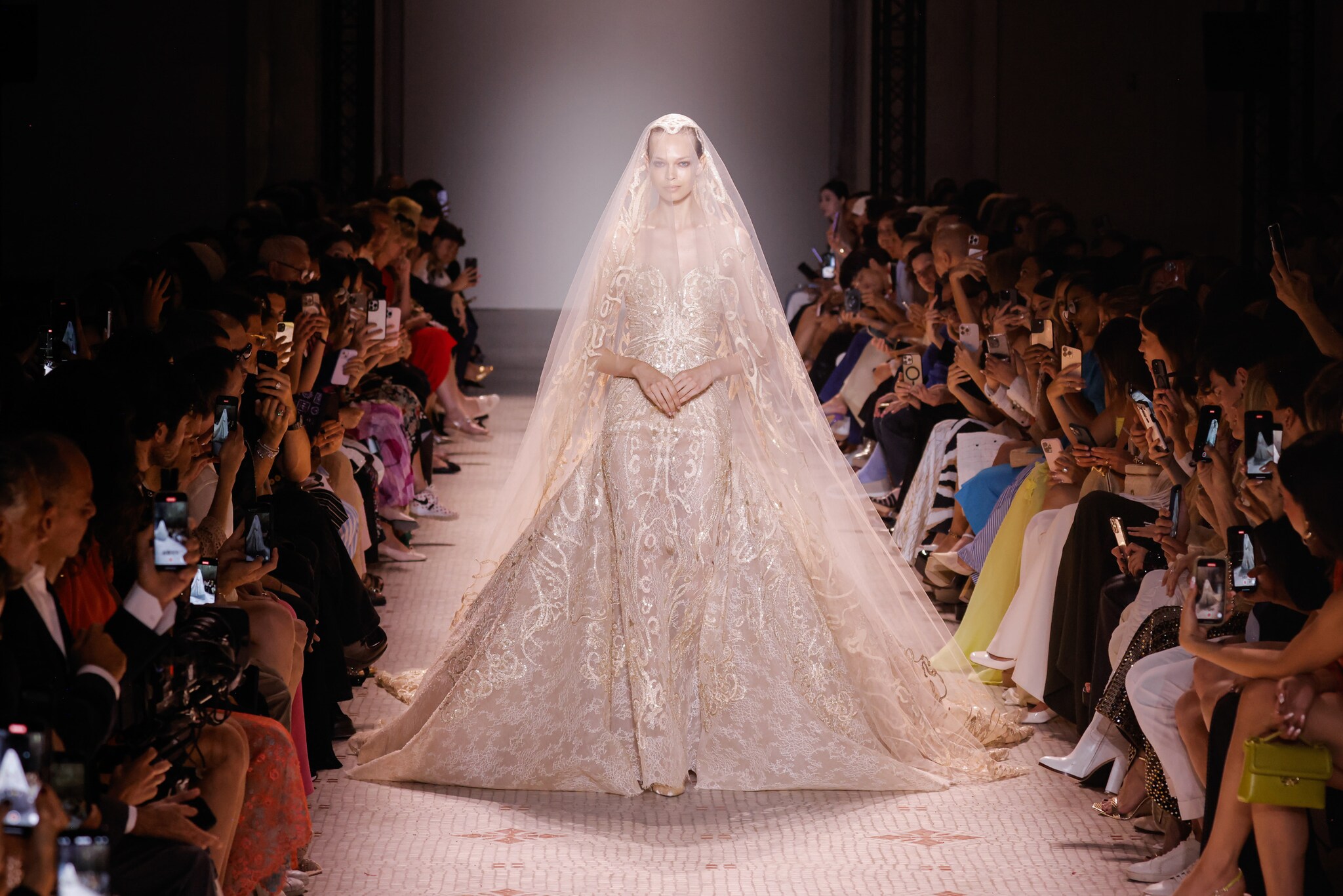 Ein Model in einem langen bestickten Kleid mit Schleier auf dem Laufsteg bei einer Modenschau mit Publikum.