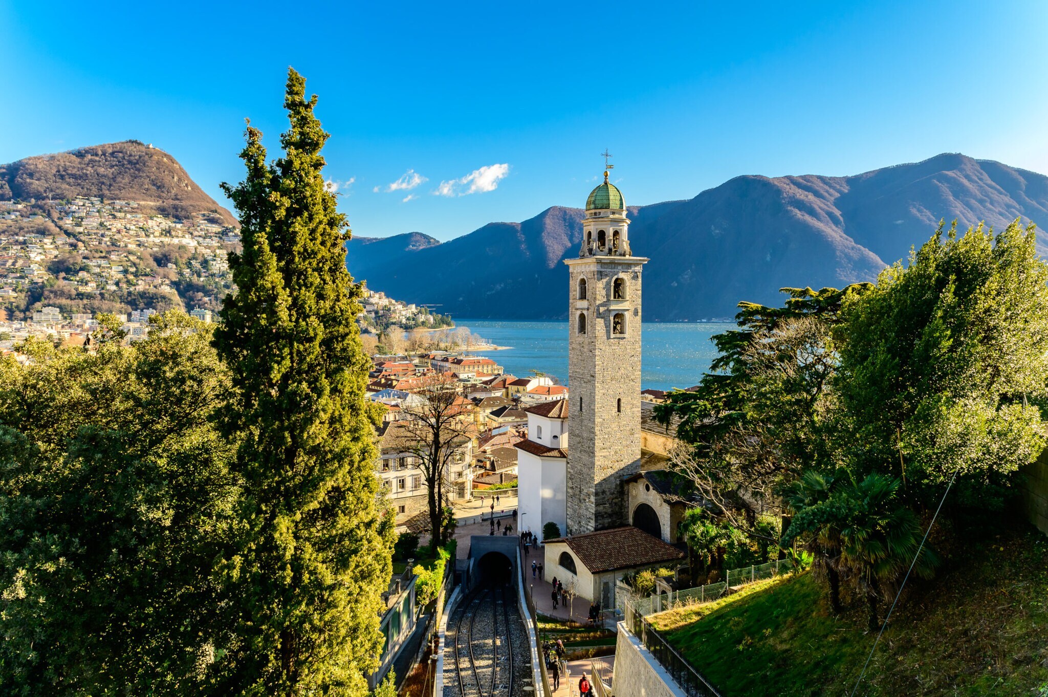 Stadtpanorama von Merano mit Zypressen und Glockenturm an einem See vor Bergpanorama.