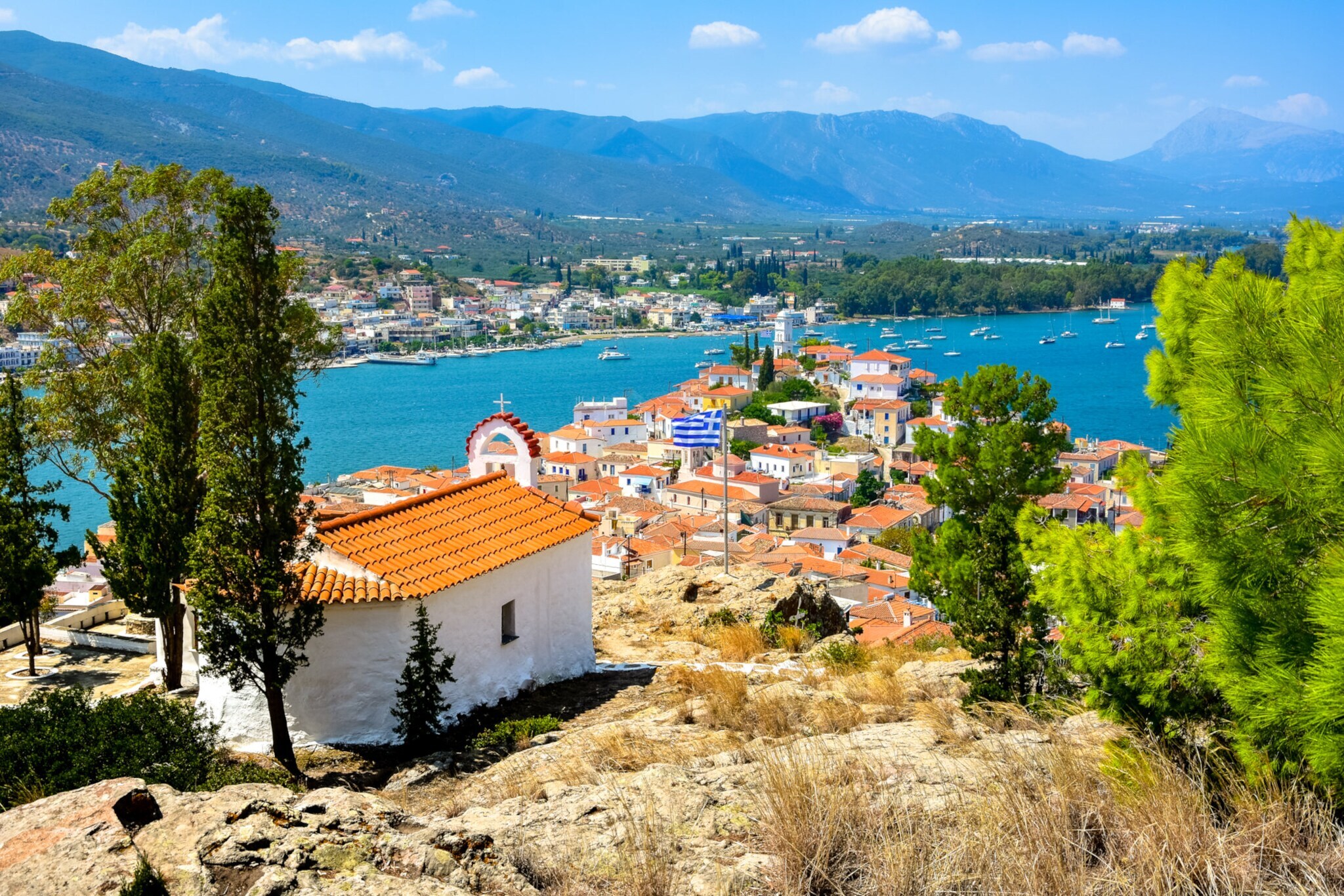 Blick auf einen mediterranen Ort von einem Felsen mit griechischer Flagge vor Küstenpanorama.