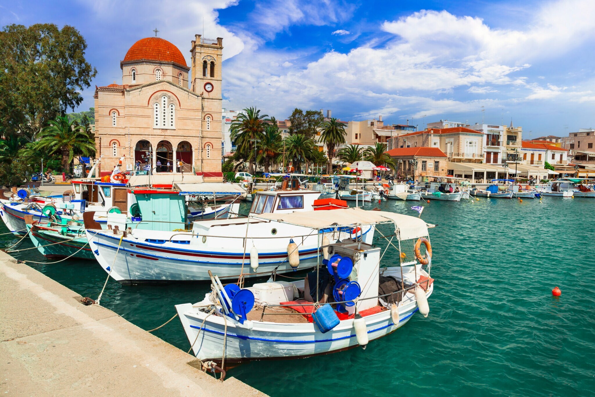 Mediterraner Ort mit Kirche an Hafenpromenade mit Fischerbooten im Vordergrund.