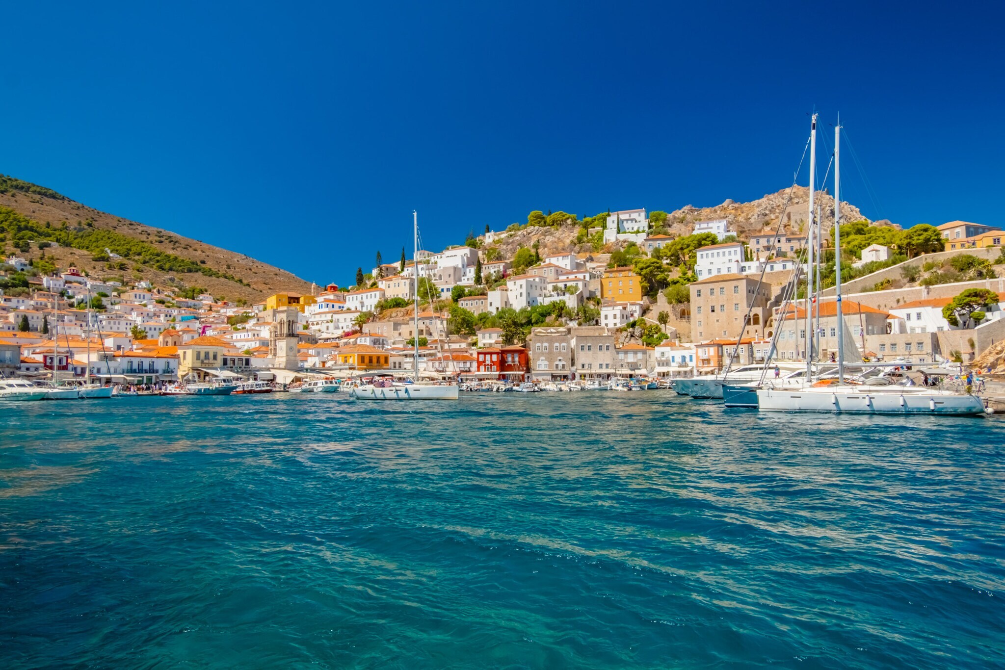 Mediterrane Ortschaft in Hügellage mit Hafenpromenade, im Vordergrund türkisblaues Meer mit Booten.