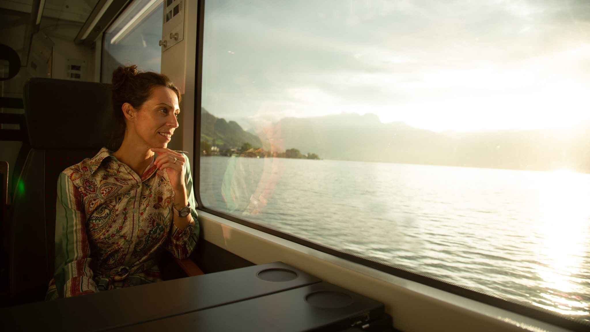 Eine Frau sitzt in einem Zug und blickt durch ein Panoramafenster auf einen See. Eine Frau sitzt in einem Zug und blickt durch ein Panoramafenster auf einen See.