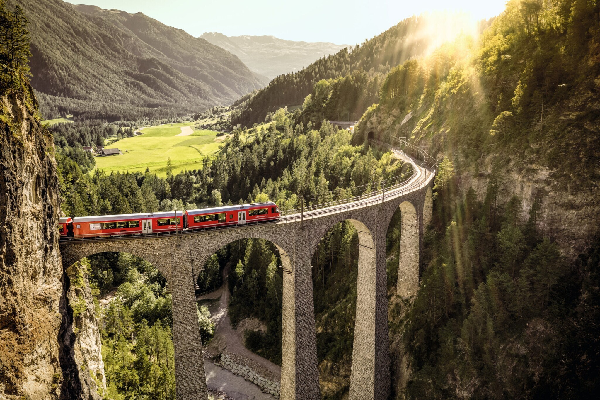 Ein roter Zug fährt auf einem Viadukt durch eine grüne Berglandschaft.