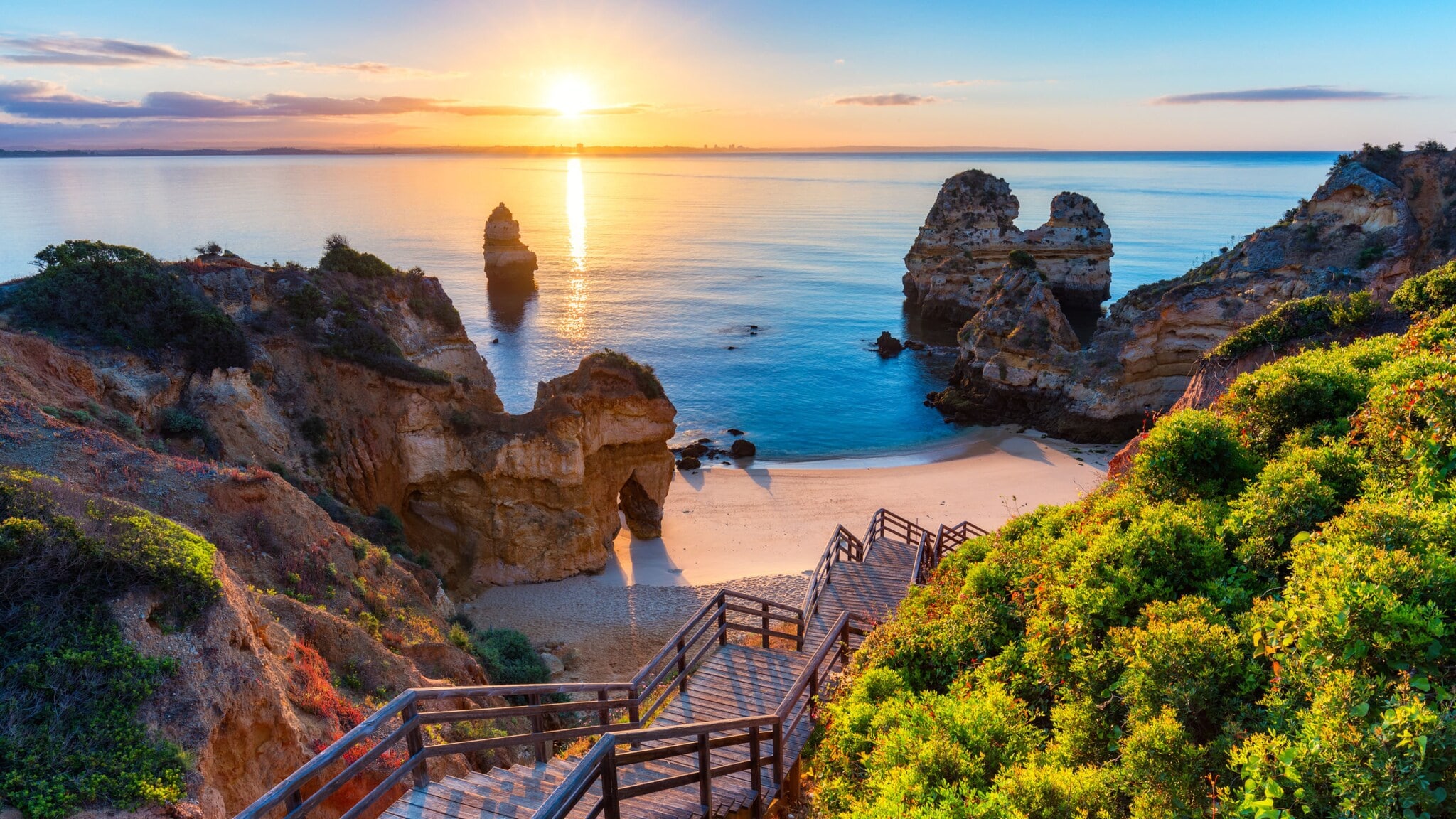 Der Strand Praia do Camilo in Portugal bei Sonnenuntergang, zu dem über Felsen eine Holztreppe nach unten führt. Der Strand Praia do Camilo in Portugal bei Sonnenuntergang, zu dem über Felsen eine Holztreppe nach unten führt.