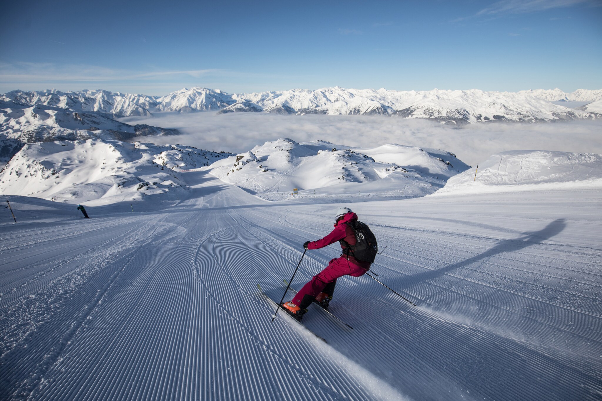 Rückansicht einer Skifahrerin auf einer breiten Piste vor schneebedeckten Gipfeln.