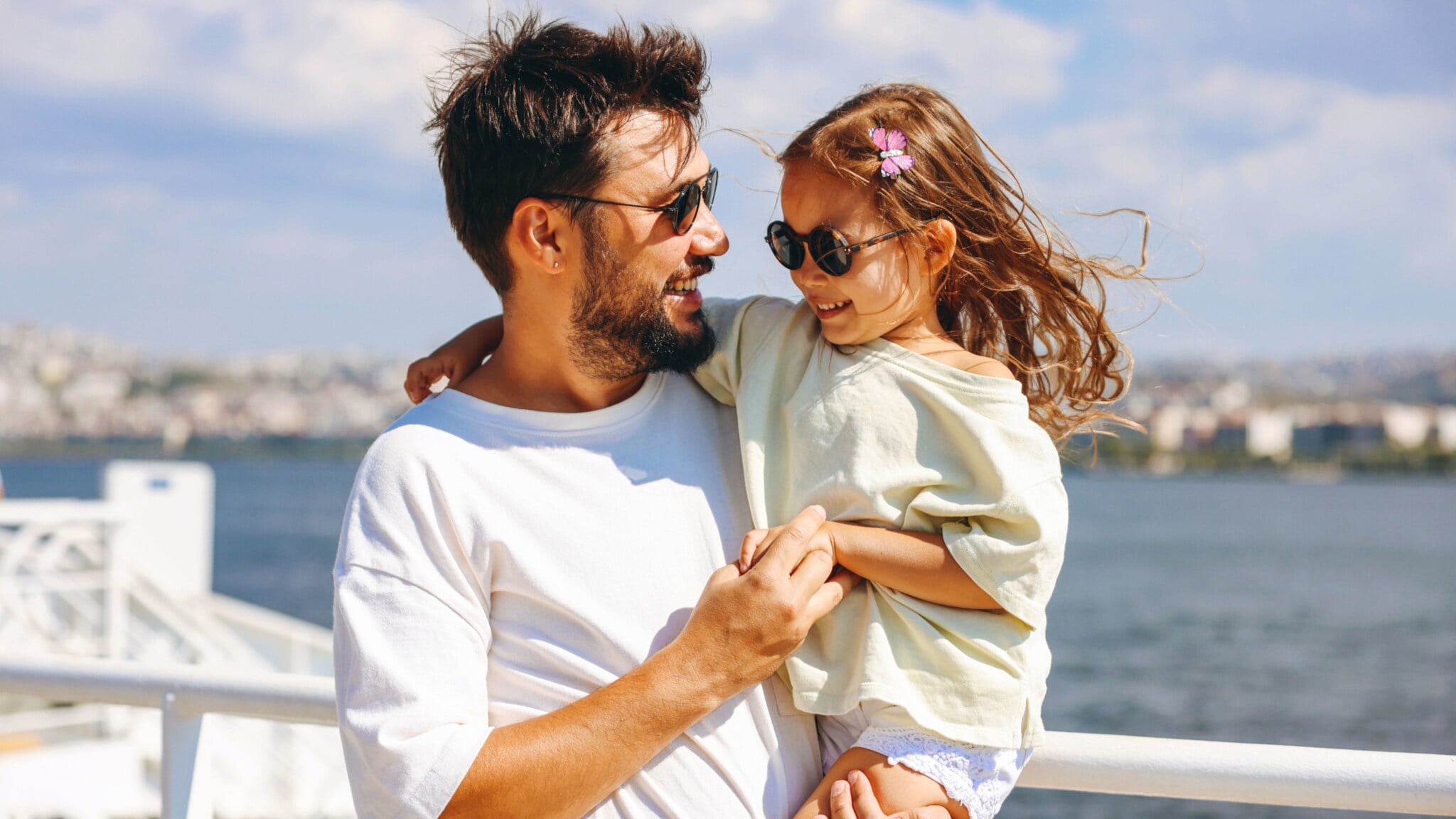 Ein lächelnder Mann mit Sonnbrille mit einem fröhlichen Mädchen mit Sonnenbrille auf dem Arm auf dem Außendeck eines Schiffes. Ein lächelnder Mann mit Sonnbrille mit einem fröhlichen Mädchen mit Sonnenbrille auf dem Arm auf dem Außendeck eines Schiffes.