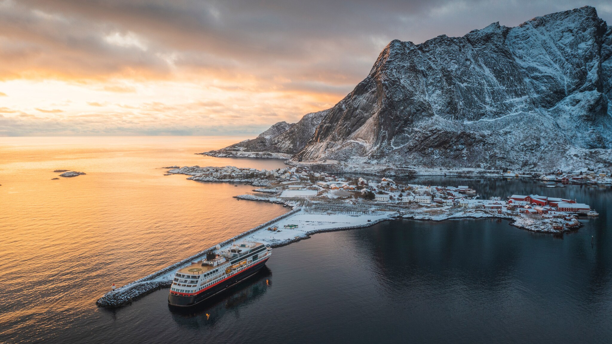 Luftaufnahme von einem Kreuzfahrtschiff, vor Anker an einer schneebedeckten Ortschaft vor kargem Bergpanorama bei Sonnenuntergang.