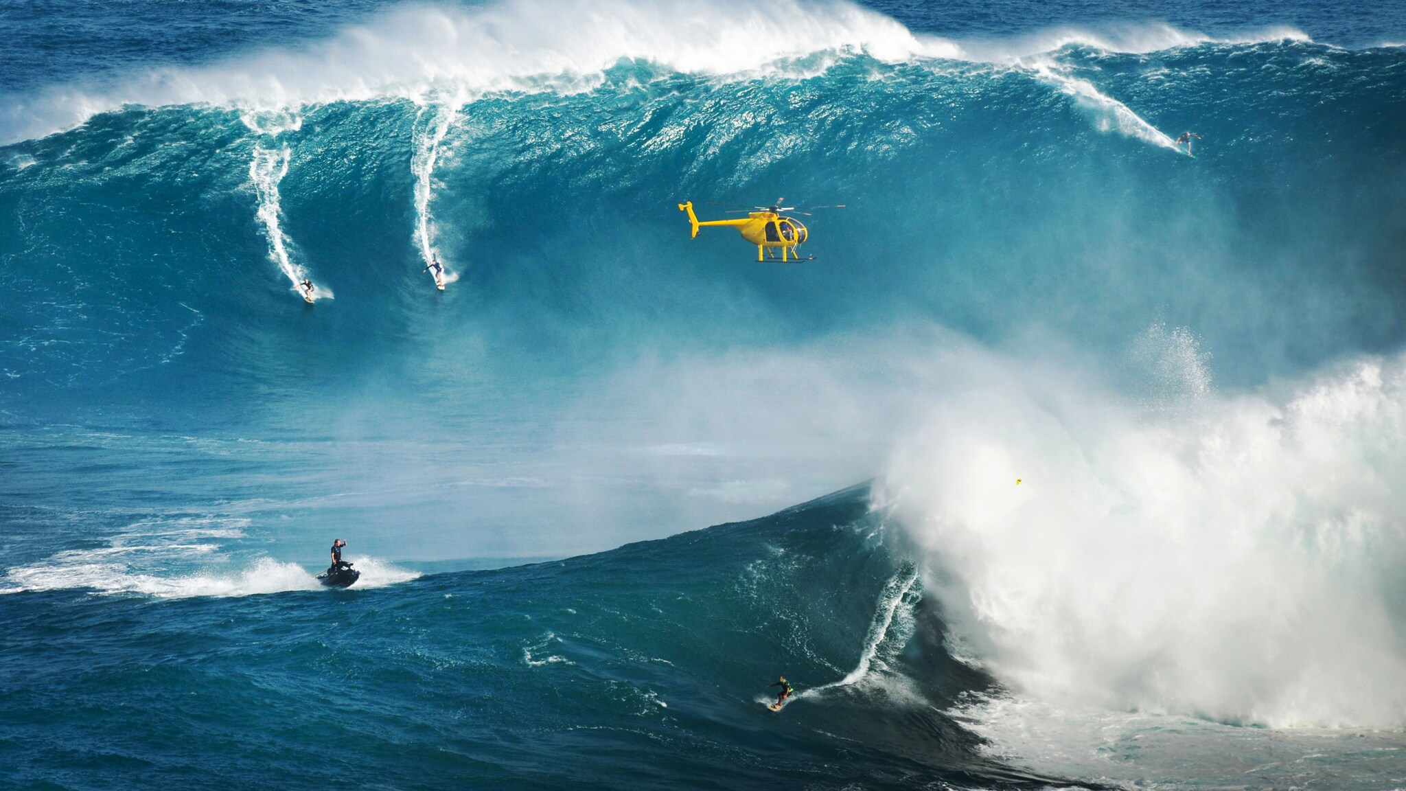 Surfende und ein Jetski auf gigantischen Wellen im Meer, über ihnen fliegt ein gelber Helikopter.