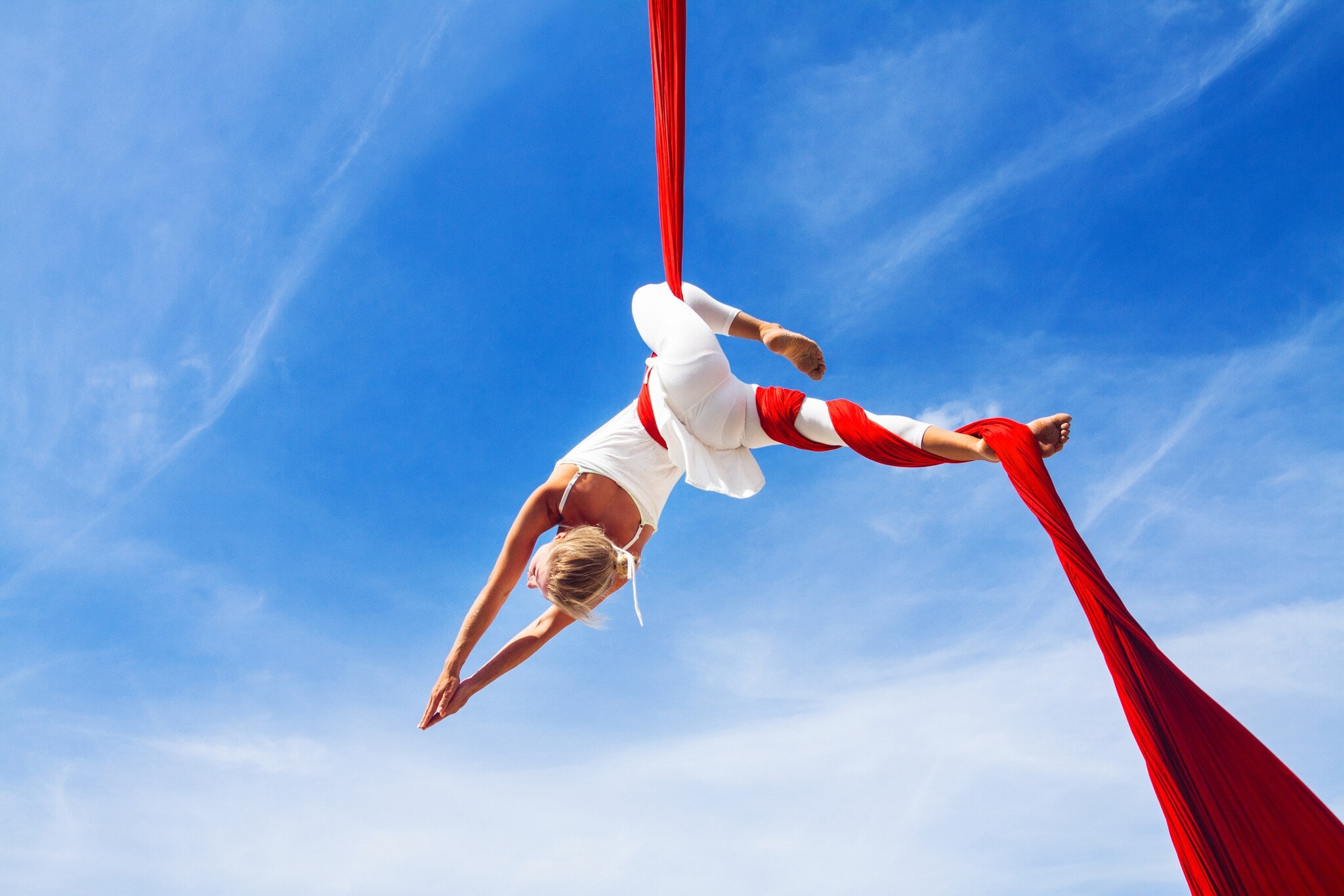 Untersicht einer Frau in weißer Kleidung in schwebender Yoga-Pose in einem roten Tuch vor blauem Himmel. Untersicht einer Frau in weißer Kleidung in schwebender Yoga-Pose in einem roten Tuch vor blauem Himmel.