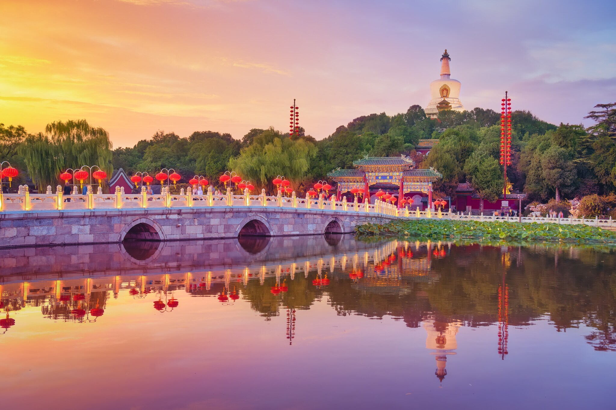 Landschaftsgarten mit einer mit roten Lampions dekorierten Brücke vor einer Tempelanlage.
