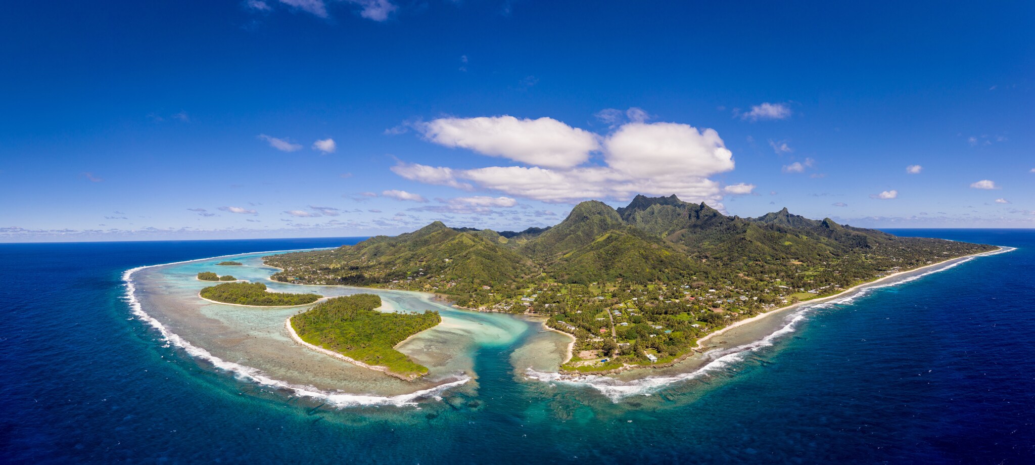 Luftaufnahme der Südseeinsel Rarotonga mit grüner Vulkanlandschaft und türkisblauer Lagune, umgeben von blauem Meer.