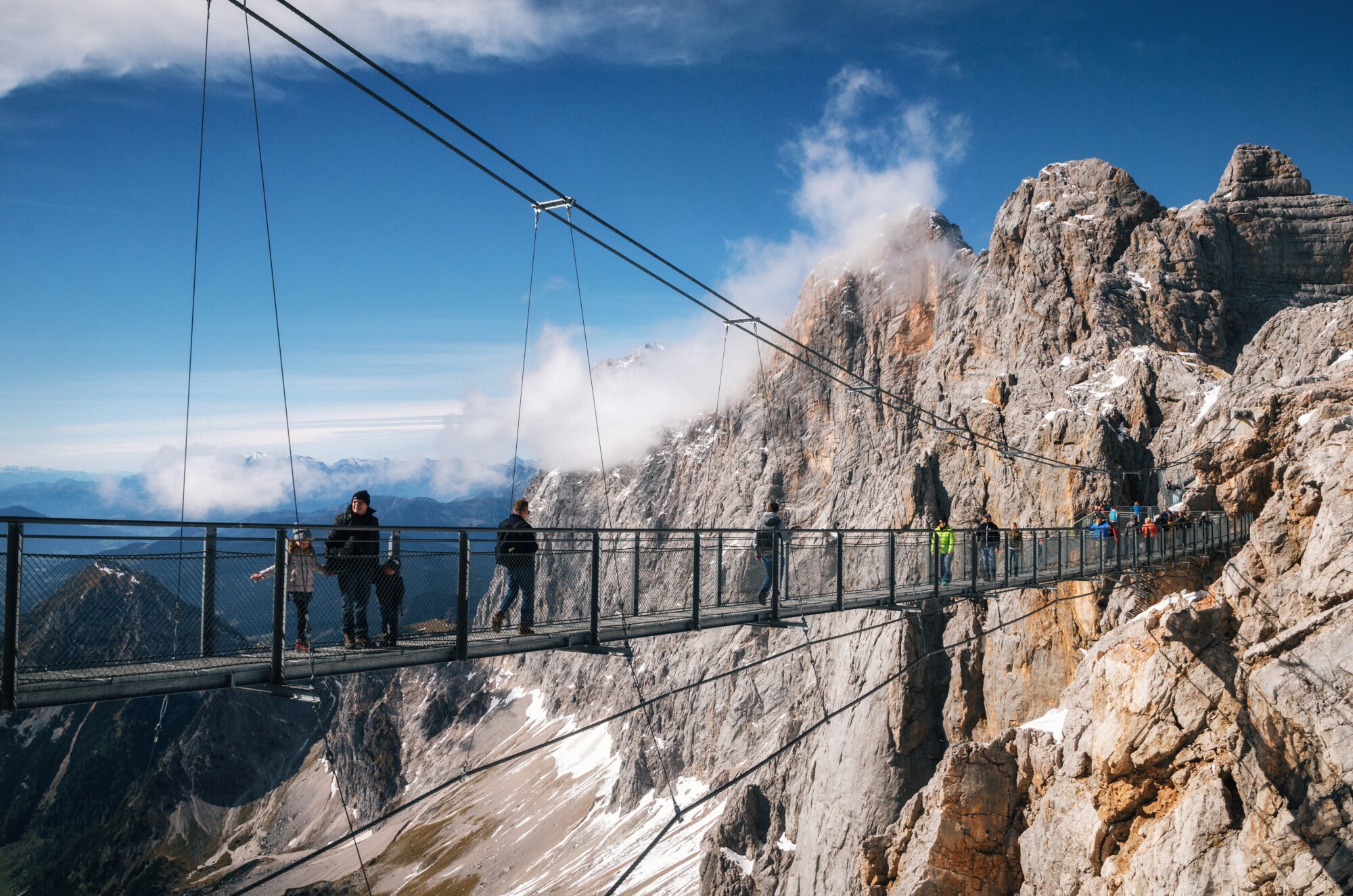 Personen auf einer Hängebrücke vor einer Felswand im Gebirge.