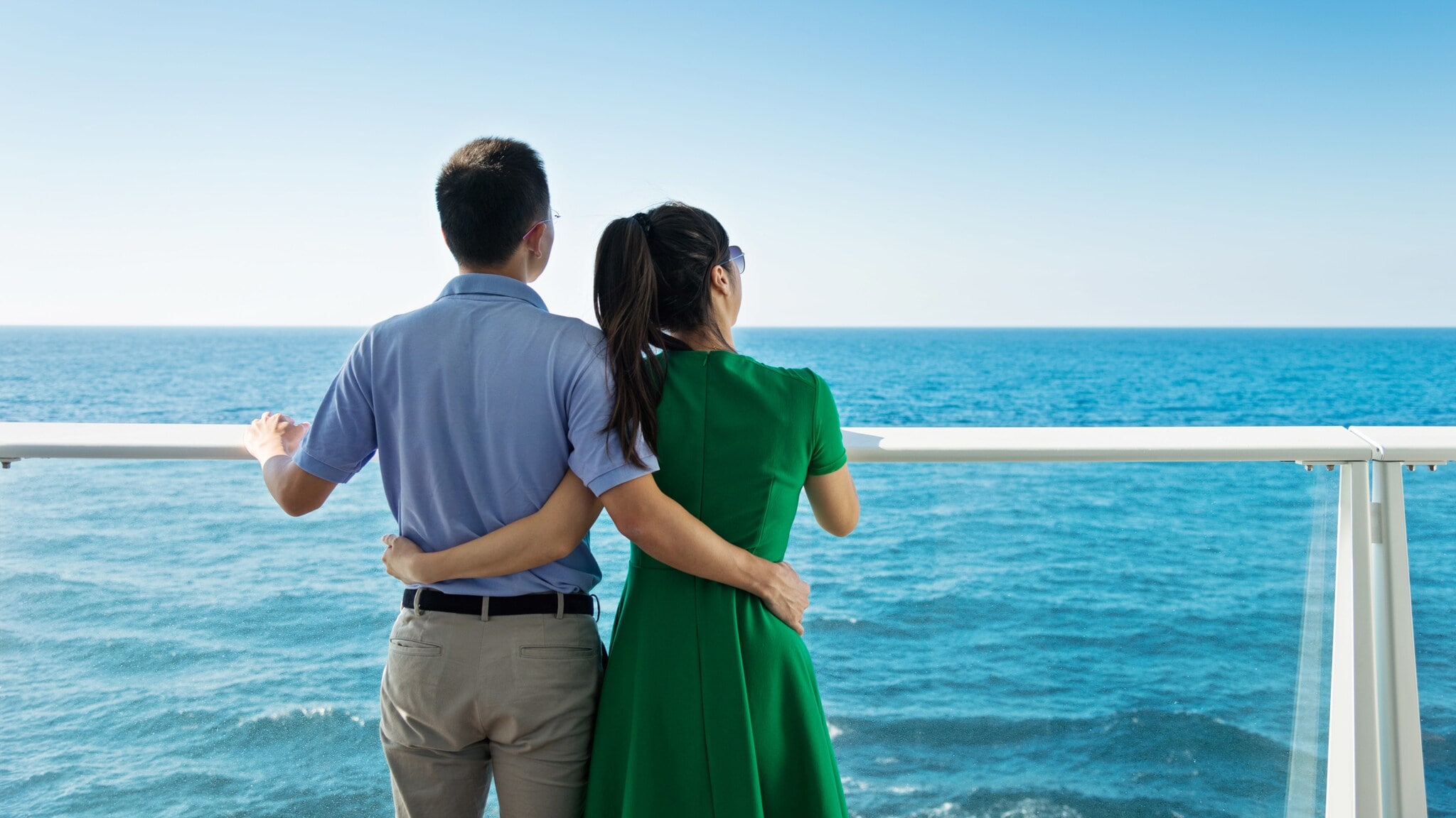 Rückansicht eines sich umarmenden Paars an einer Reling vor Meerespanorama, sie trägt ein grünes Kleid, er Hemd mit Stoffhose. Rückansicht eines sich umarmenden Paars an einer Reling vor Meerespanorama, sie trägt ein grünes Kleid, er Hemd mit Stoffhose.