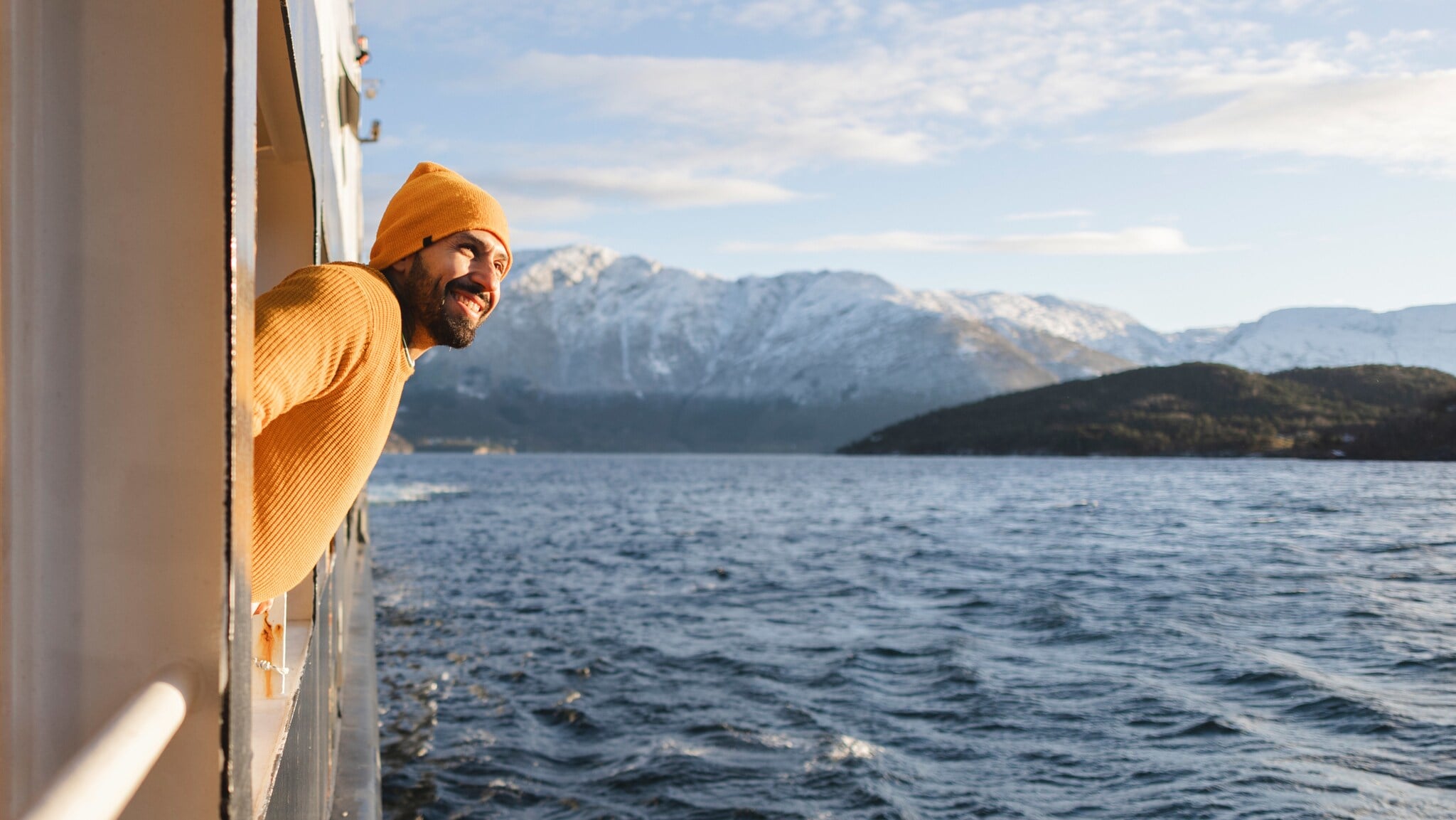 Ein lächelnder Mann mit gelbem Pullover und Mütze schaut von einem Schiff aufs Wasser vor schneebedeckter Berglandschaft. Ein lächelnder Mann mit gelbem Pullover und Mütze schaut von einem Schiff aufs Wasser vor schneebedeckter Berglandschaft.