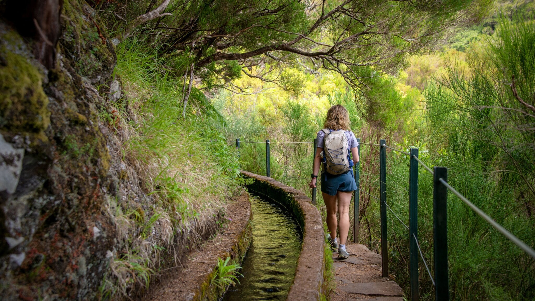 Rückansicht einer Frau auf einem Wanderweg auf Madeira, der entlang eines Bewässerungskanals verläuft. Rückansicht einer Frau auf einem Wanderweg auf Madeira, der entlang eines Bewässerungskanals verläuft.
