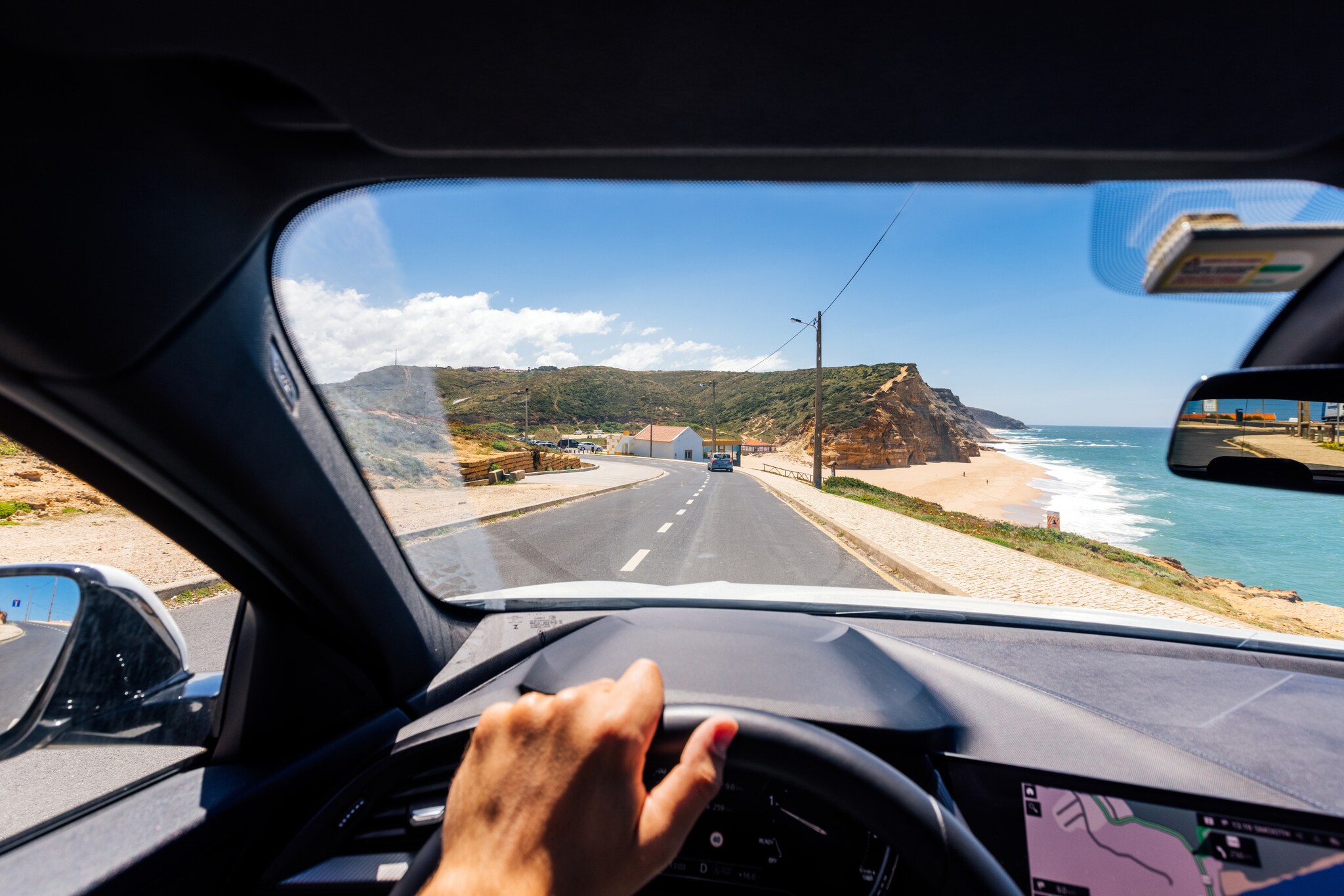 Blick über ein Lenkrad durch die Frontscheibe eines fahrenden Autos auf einer Straße an einer Küste mit Sandstrand bei Sonnenschein. Blick über ein Lenkrad durch die Frontscheibe eines fahrenden Autos auf einer Straße an einer Küste mit Sandstrand bei Sonnenschein.