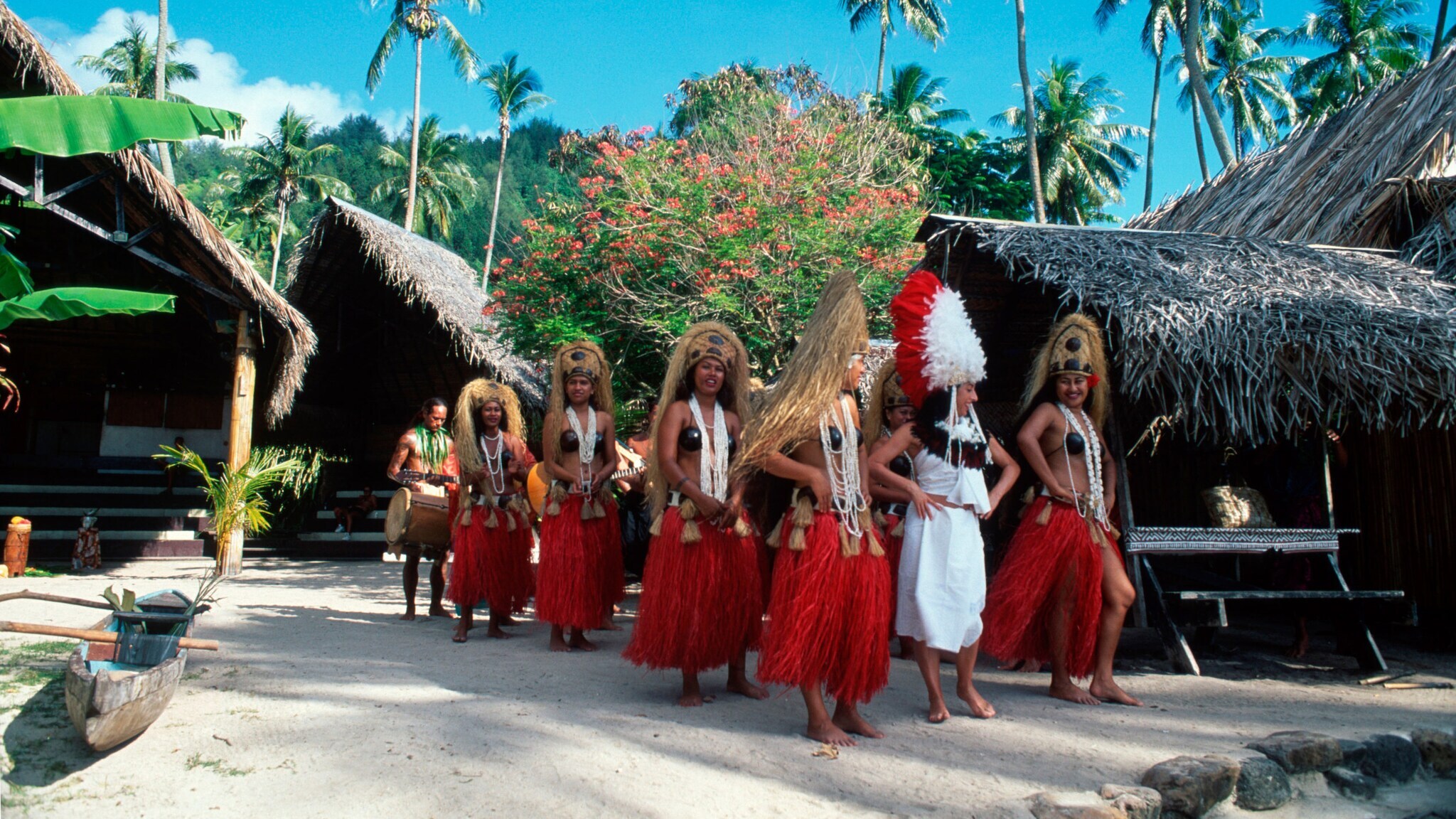 Aufgereihte Frauen mit roten Baströcken und Kopfschmuck in einem polynesischen Dorf.