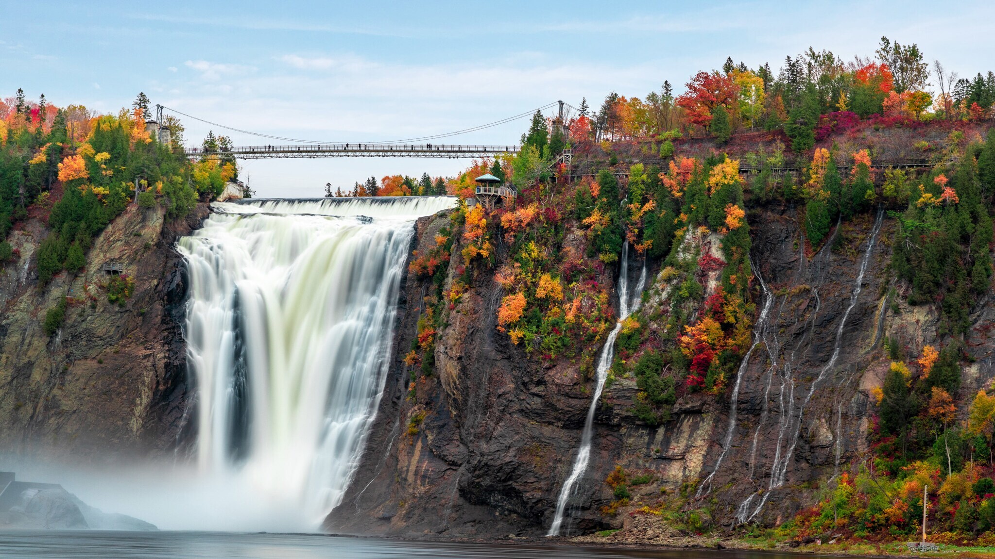 Personen auf einer Brücke über einem Wasserfall, umgeben von Bäumen mit herbstlicher Verfärbung.