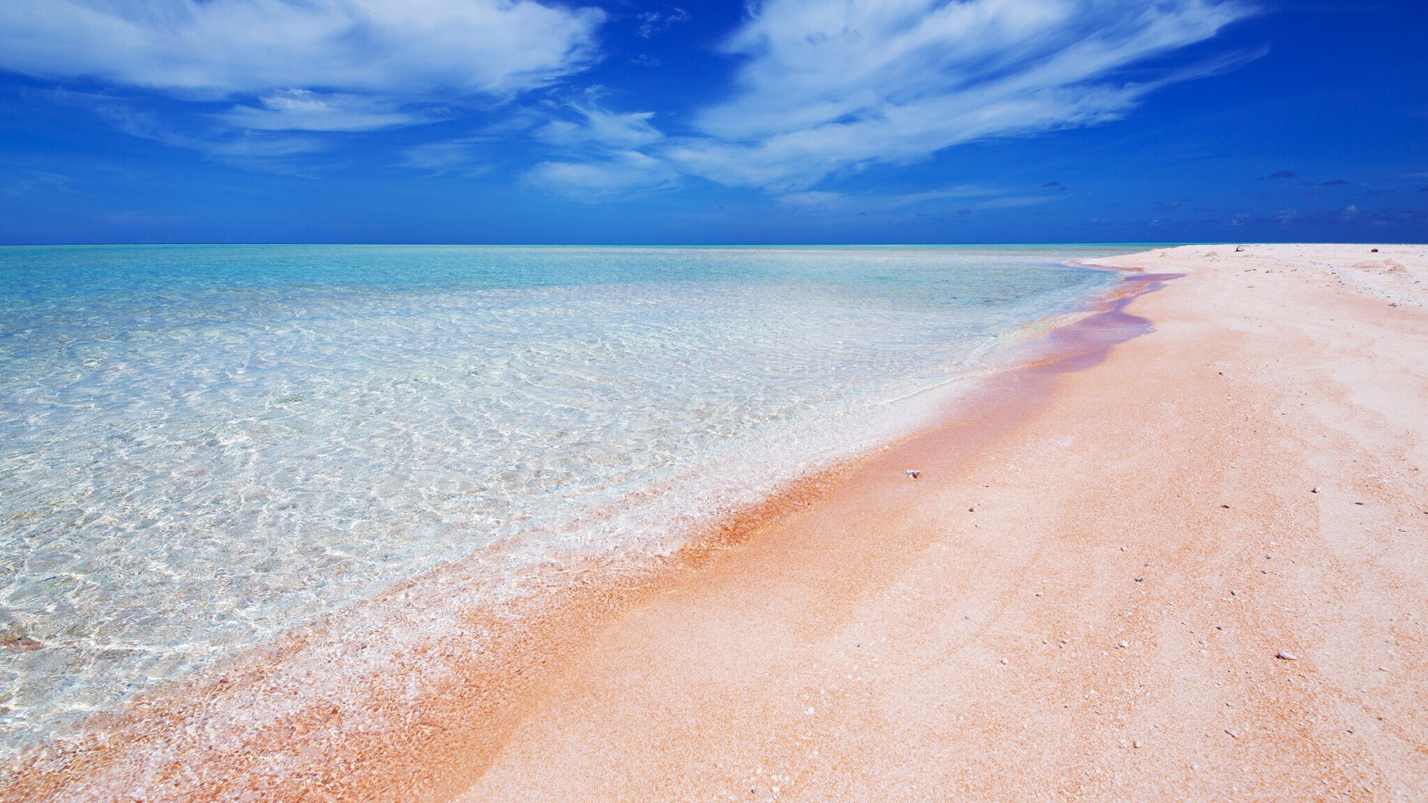 Weiter Sandstrand mit rosa Färbung an klarem Wasser unter blauem Himmel.