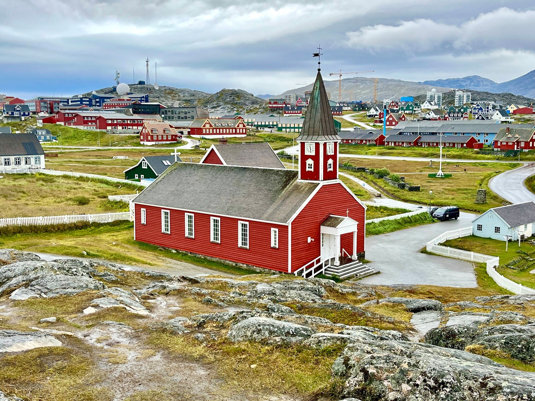 Das Bild zeigt die Kirche der grönländischen Hauptstadt Nuuk. Rund um die mit rot angestrichenem Holz verkleidete Kirche sind grüne Wiesen und kleine Gebäude zu sehen. Das Bild zeigt die Kirche der grönländischen Hauptstadt Nuuk. Rund um die mit rot angestrichenem Holz verkleidete Kirche sind grüne Wiesen und kleine Gebäude zu sehen.