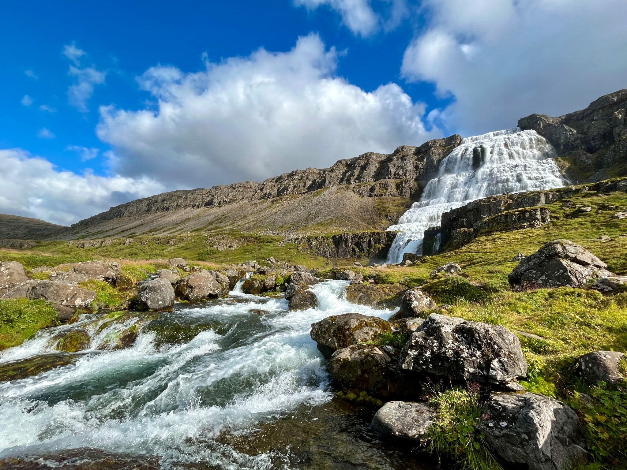 Das Bild zeigt im Hintergrund den Dynjandi-Wasserfall im Nord-Westen von Island. Im Vordergrund ist das über Felsen sprudelnde Wasser zwischen grünen Wiesen zu sehen. Das Bild zeigt im Hintergrund den Dynjandi-Wasserfall im Nord-Westen von Island. Im Vordergrund ist das über Felsen sprudelnde Wasser zwischen grünen Wiesen zu sehen.
