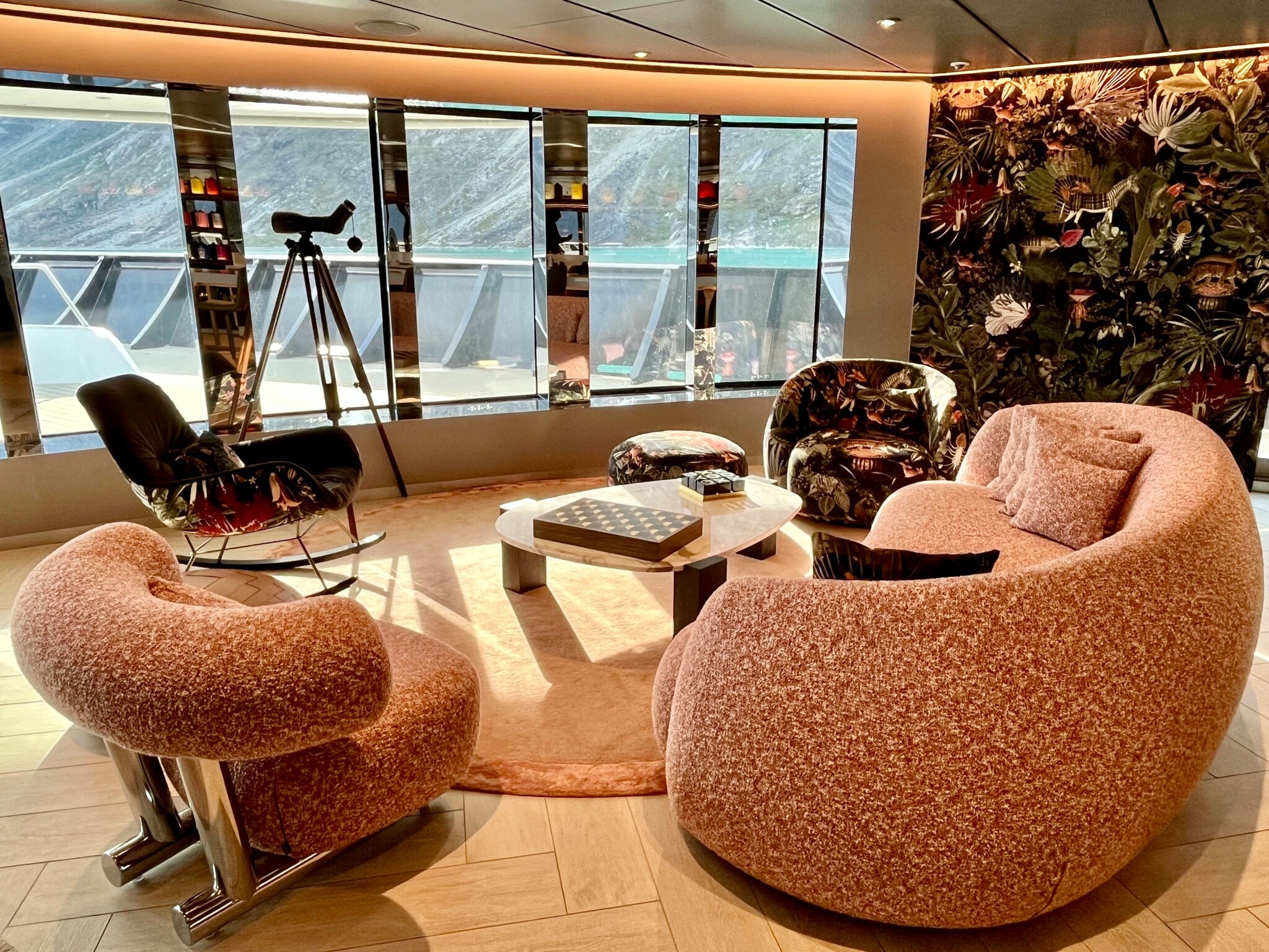 Das Bild zeigt eine Sitzgruppe in der Observation Lounge der Scenic Eclipse II. Auf dem Tisch liegt ein Schachspiel. Im Hintergrund ist ein Fernglas zu sehen. Bodentiefe Panoramafenster ermöglichen den Blick auf die Berge in der Umgebung. Das Bild zeigt eine Sitzgruppe in der Observation Lounge der Scenic Eclipse II. Auf dem Tisch liegt ein Schachspiel. Im Hintergrund ist ein Fernglas zu sehen. Bodentiefe Panoramafenster ermöglichen den Blick auf die Berge in der Umgebung.