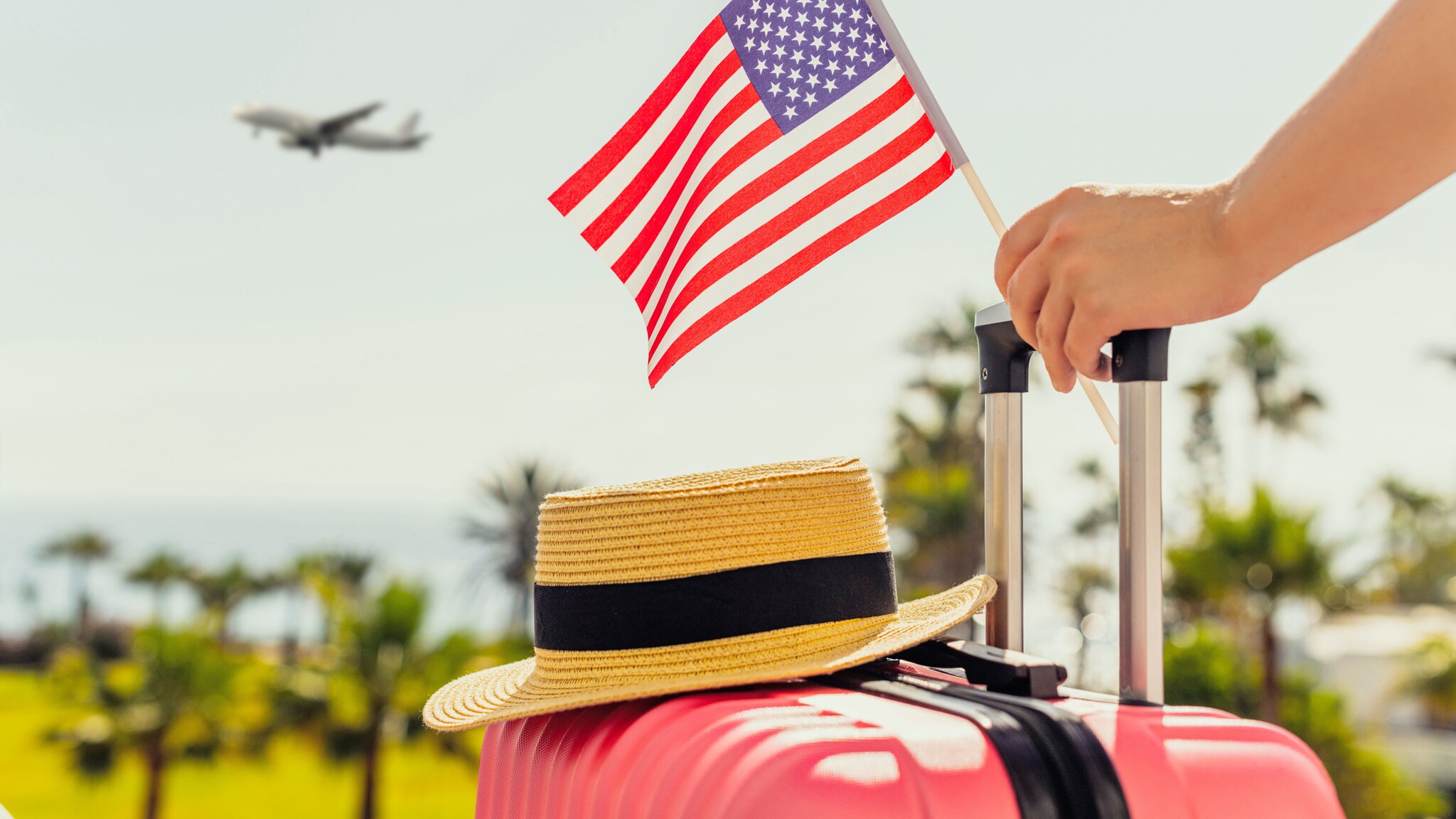 Ein Strohhut auf einem pinken Hartschalenkoffer, eine Hand am Griff hält eine kleine US-amerikanische Flagge, im Hintergrund Plamen und ein Flugzeug am Himmel.