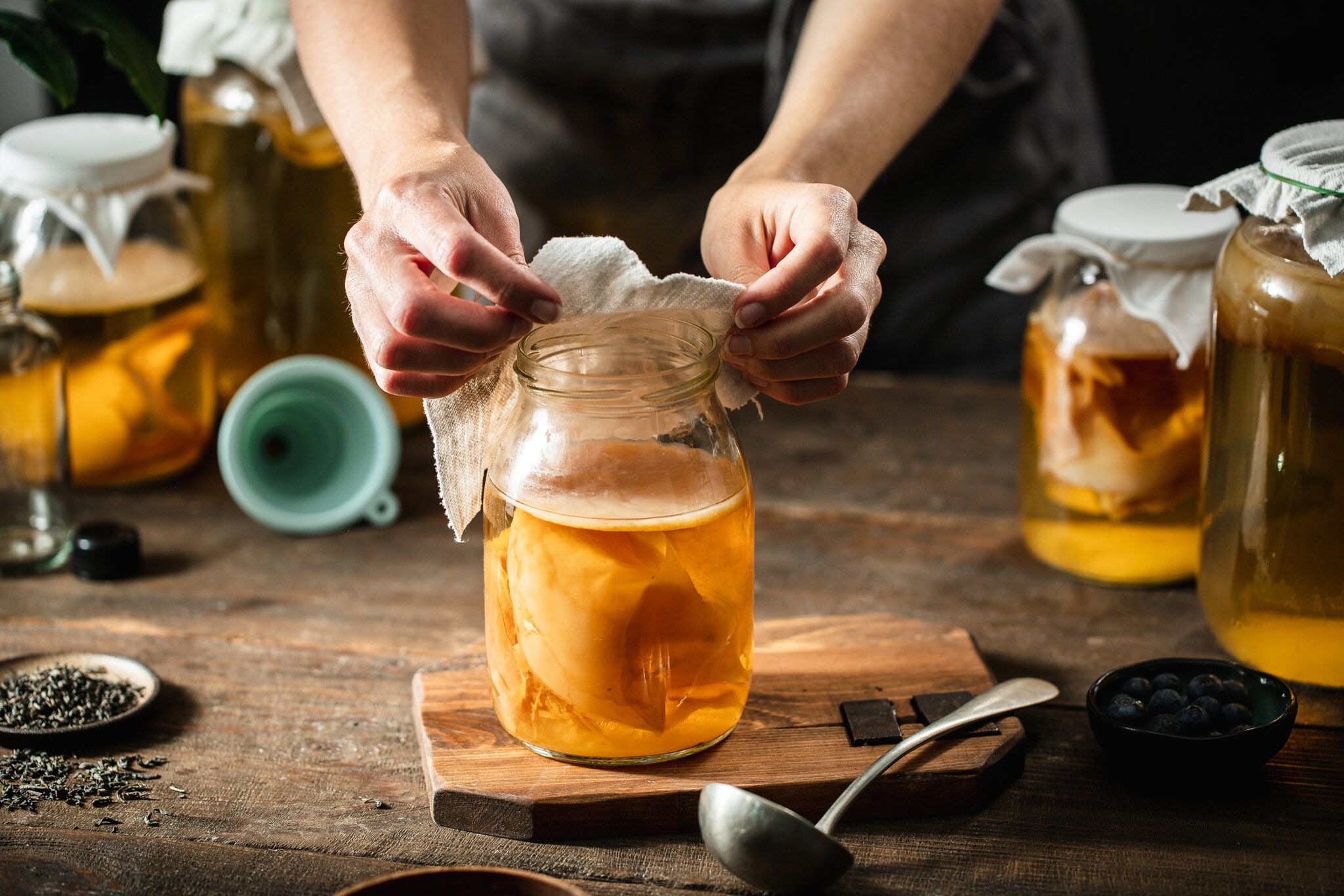 Zwei Hände bedecken mit einem Stofftuch ein Einmachglas mit Kombucha-Getränk samt Teepilz auf einem Holztisch, daneben weitere befüllte Einmachgläser.