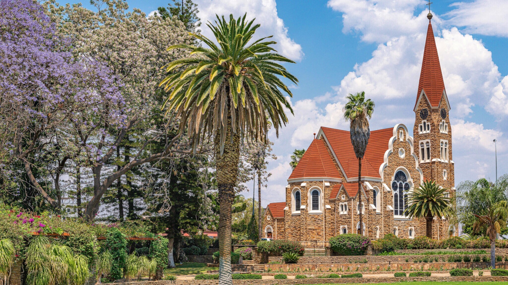 Kirche im neoromanischen Stil, im Vordergrund eine Gartenanlage mit Palmen.