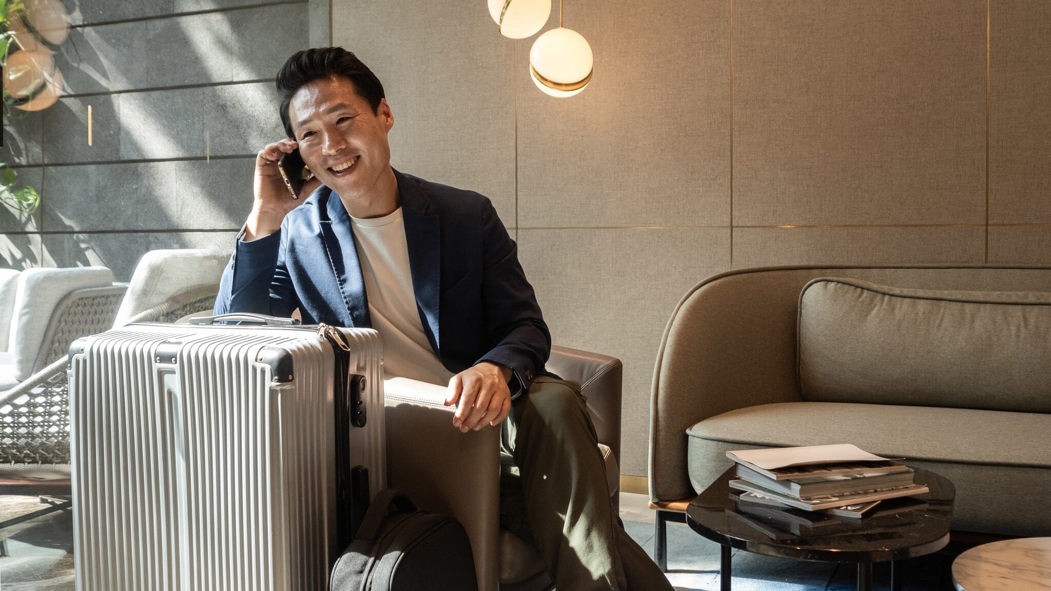 Ein fröhlicher asiatischer Geschäftsmann sitzt im Sessel einer modernen Flughafen-Lounge und telefoniert an seinem Smartphone, neben ihm steht ein großer Reisekoffer. Ein fröhlicher asiatischer Geschäftsmann sitzt im Sessel einer modernen Flughafen-Lounge und telefoniert an seinem Smartphone, neben ihm steht ein großer Reisekoffer.