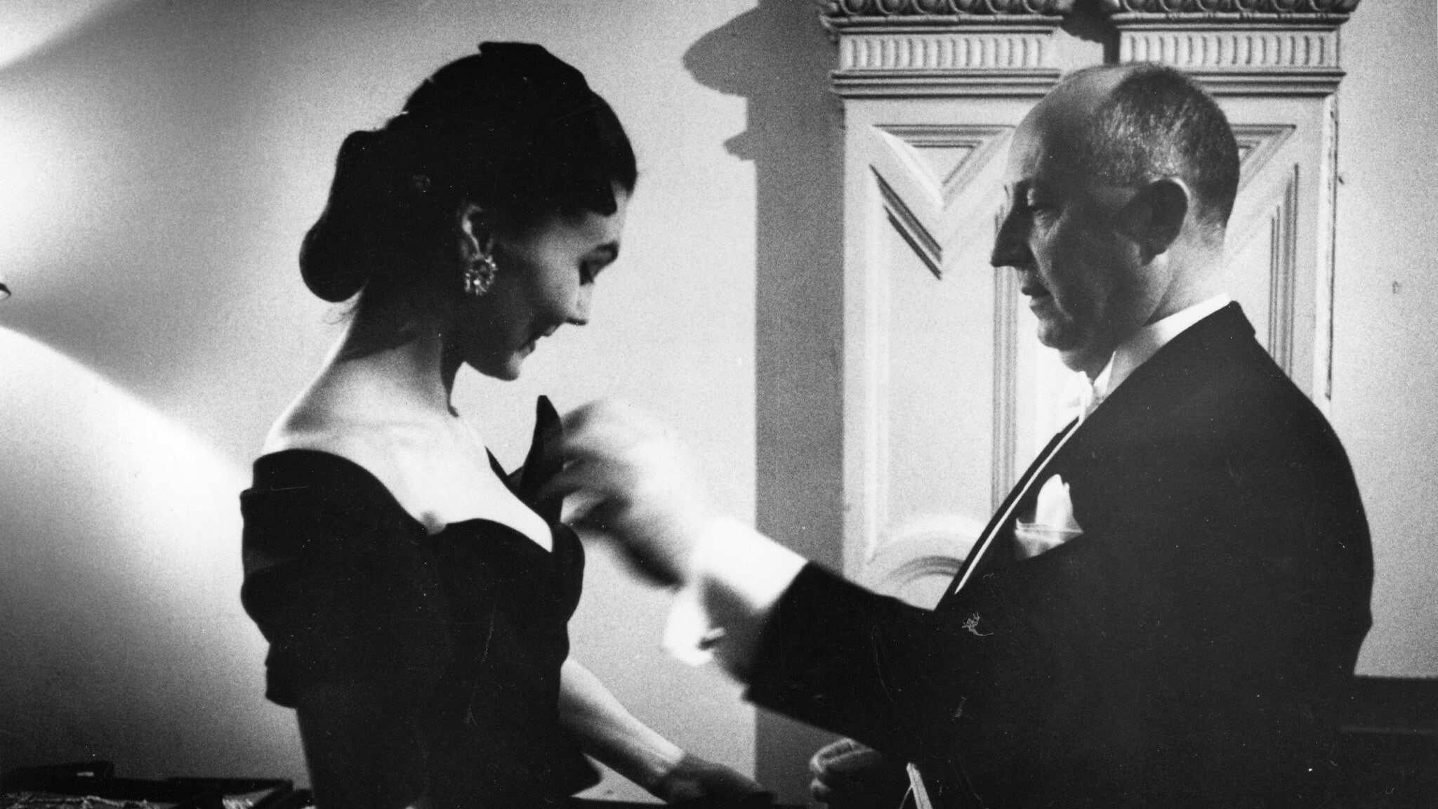 Profilansicht von Christian Dior in einem schwarzen Anzug, der etwas an einem schwarzen Abendkleid eines weiblichen Models stylt, Schwarzweißfoto. Profilansicht von Christian Dior in einem schwarzen Anzug, der etwas an einem schwarzen Abendkleid eines weiblichen Models stylt, Schwarzweißfoto.