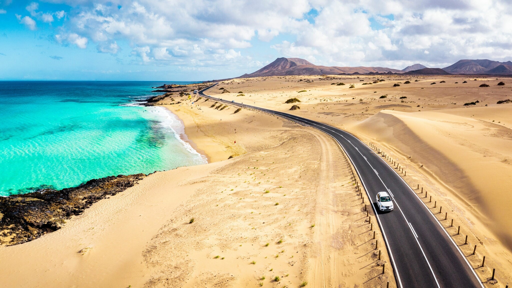Ein weißes Auto fährt auf einer Schnellstraße durch eine Wüstenlandschaft am türkisblauen Meer. Ein weißes Auto fährt auf einer Schnellstraße durch eine Wüstenlandschaft am türkisblauen Meer.
