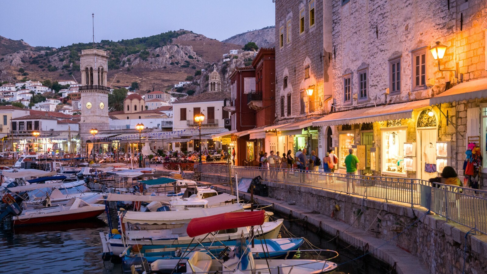 Fischerboote im Wasser entlang einer belebten Hafenpromenade in einer mediterranen Ortschaft am Abend.