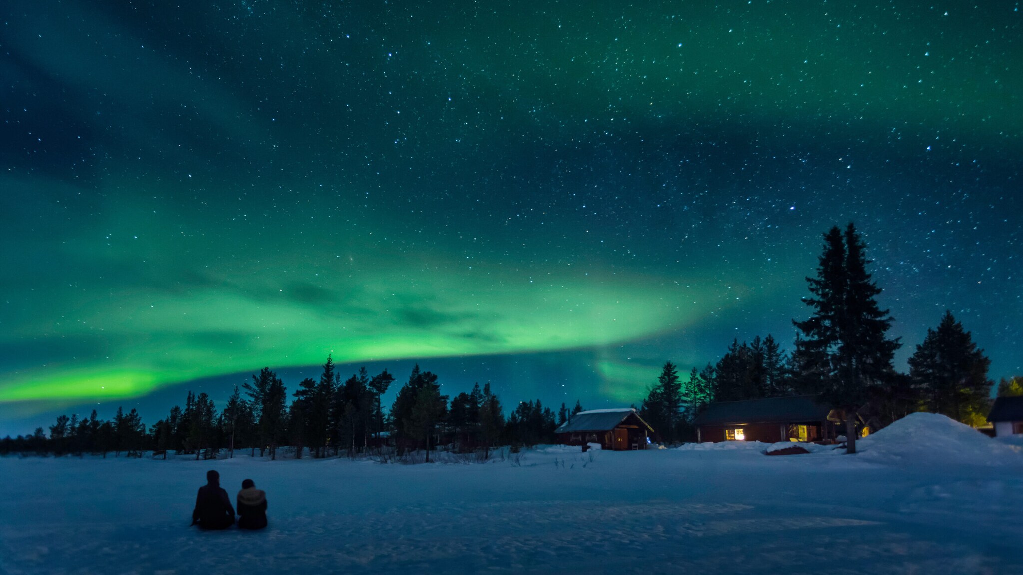 Zwei Personen sitzen im Schnee unter einem Sternenhimmel mit Polarlichtern.