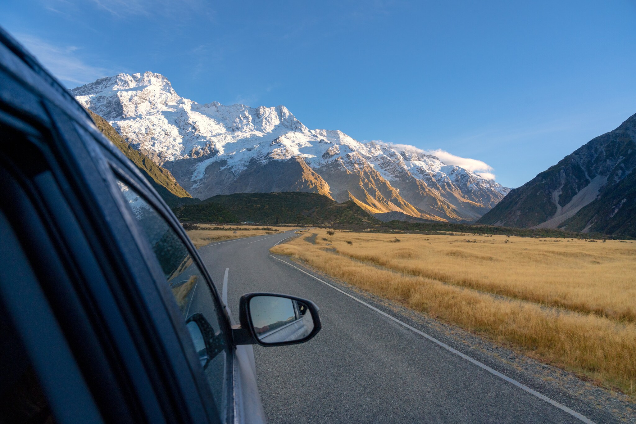 Auto mit Seitenspiegel im Anschnitt auf einer Straße vor Bergpanorama mit schneebedeckten Gipfeln.