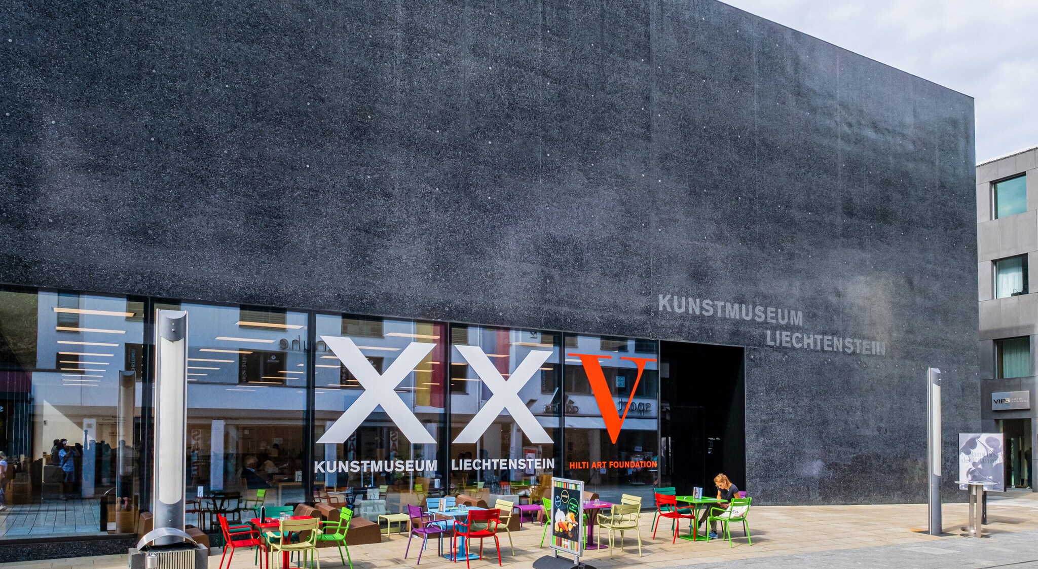 Außenansicht eines modernen, kubistischen Museumsgebäude aus schwarzem Basaltstein mit der Aufschrift Kunstmuseum Liechtenstein, im Vordergrund bunte Stühle vor einer Fensterfront. Außenansicht eines modernen, kubistischen Museumsgebäude aus schwarzem Basaltstein mit der Aufschrift Kunstmuseum Liechtenstein, im Vordergrund bunte Stühle vor einer Fensterfront.